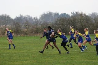 Girls playing rugby on a grassy field, with some running and others trying to tackle.