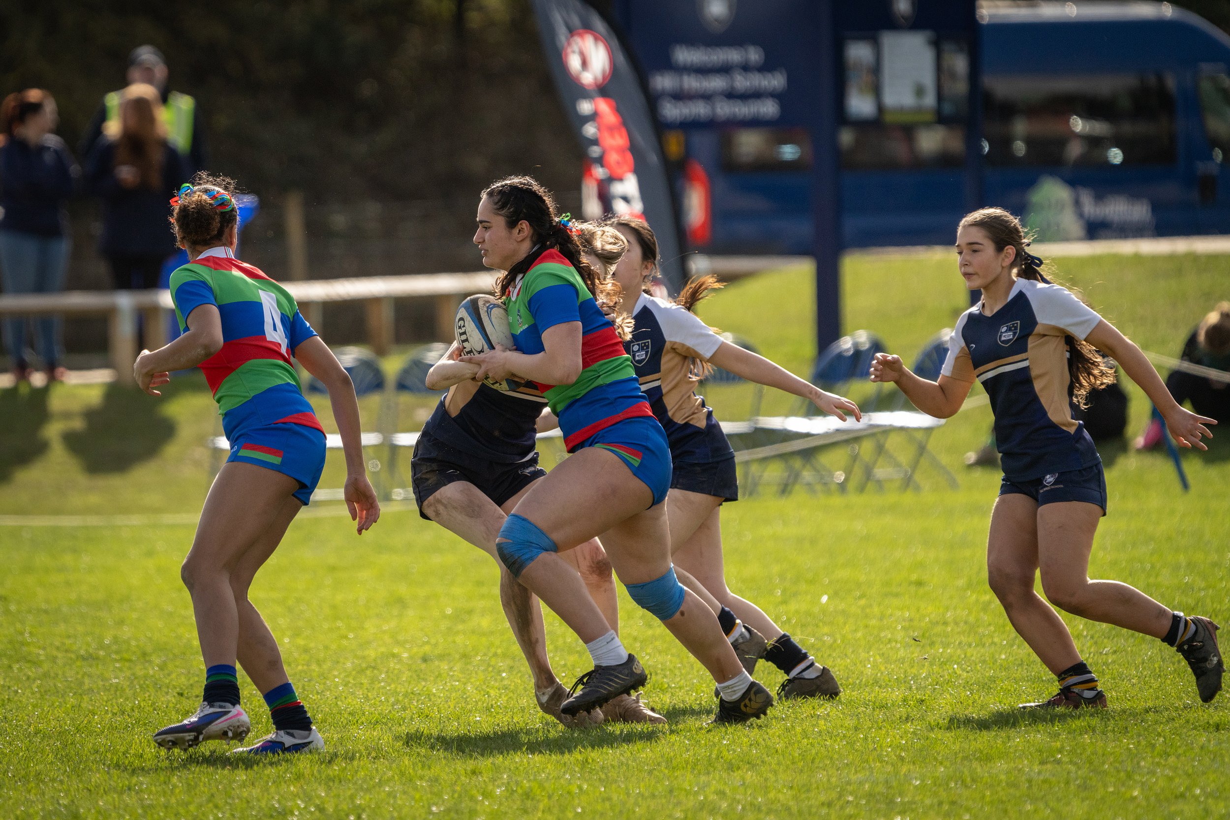 Girls playing rugby on a grass field, some wearing colorful uniforms, with a bus and spectators in the background.