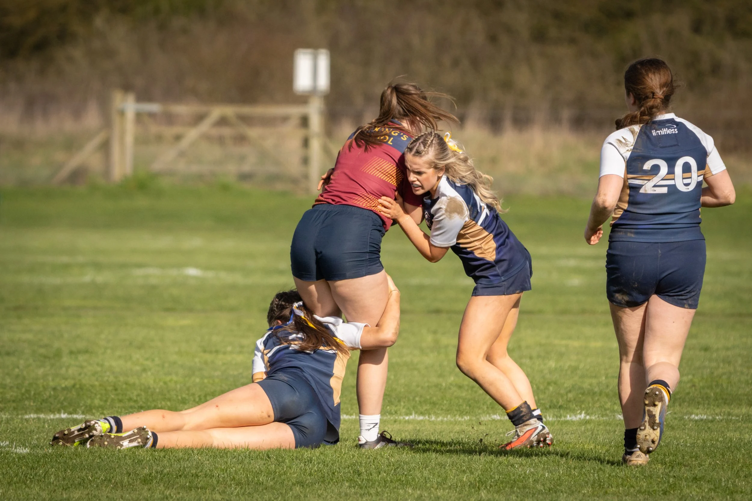 Girls playing rugby on a grassy field, with one girl on the ground and others standing nearby, during daytime.