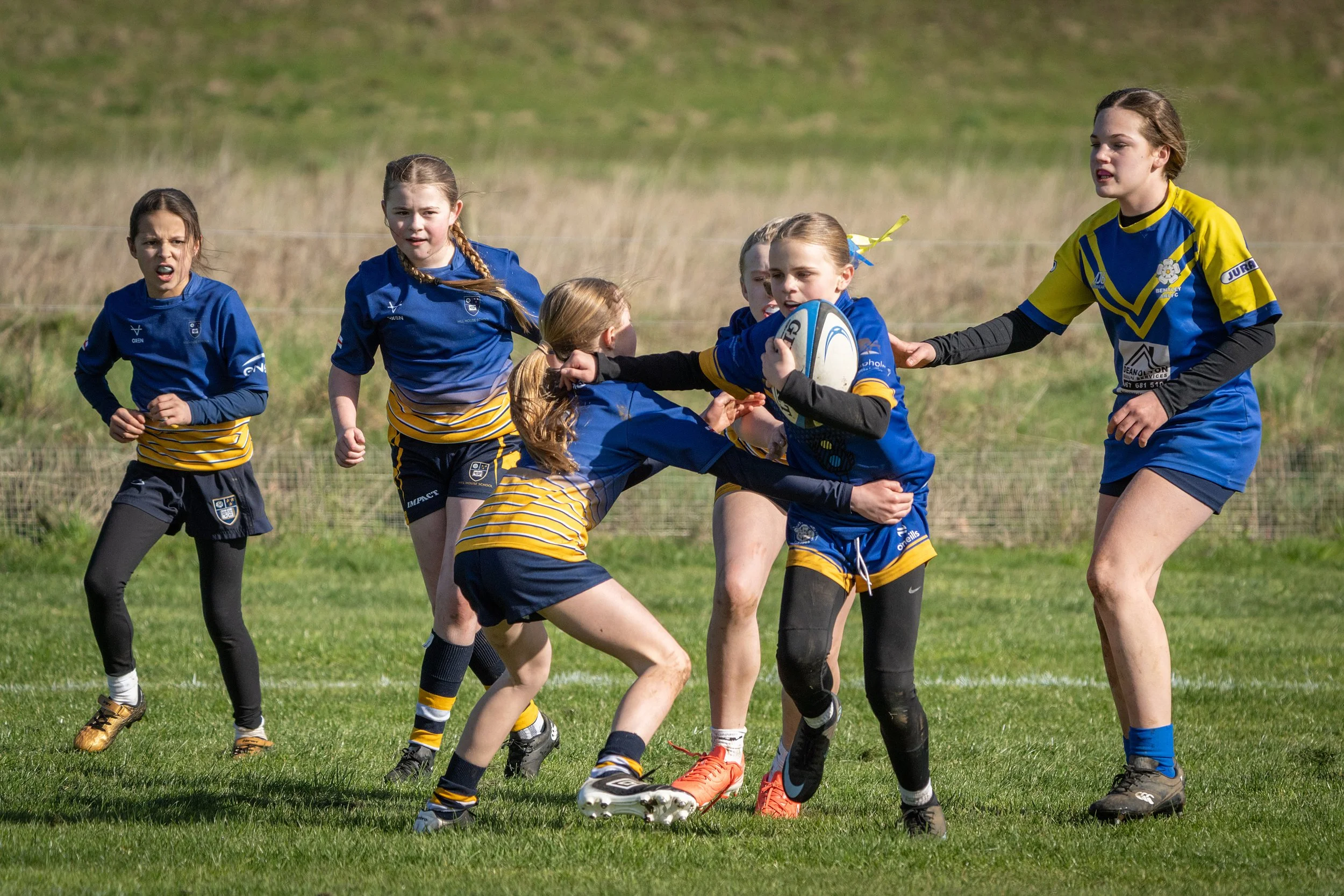 Children playing rugby on a grassy field during daytime, wearing blue and yellow uniforms.