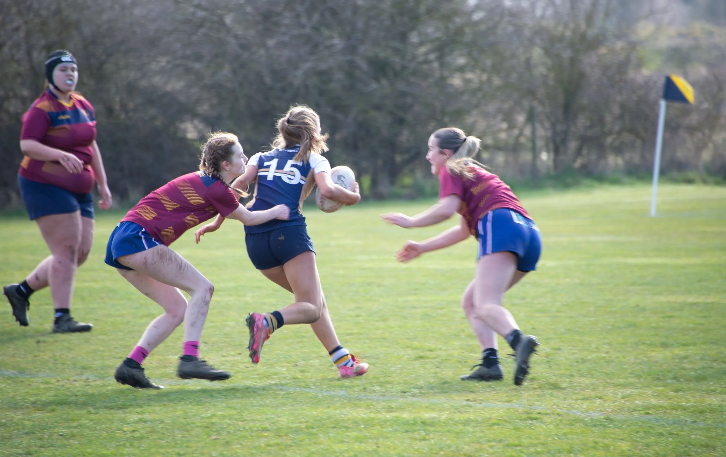 Girls in rugby uniforms playing rugby on a grassy field, with one girl holding a rugby ball and others attempting to tackle her.