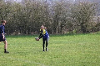 A girl holding a rugby ball on a grassy field with trees in the background, while a boy stands nearby.