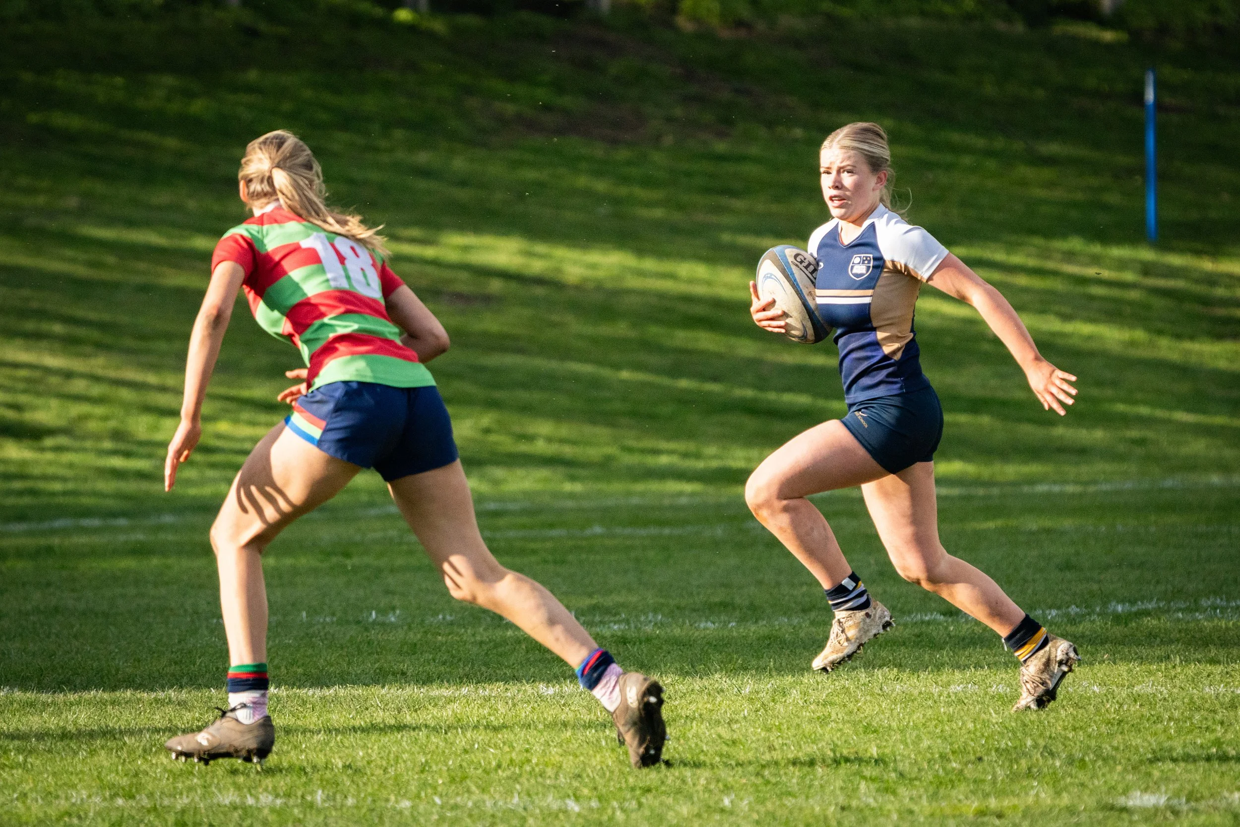 Two female rugby players running on a grassy field during a match, with one holding a rugby ball and both wearing sports uniforms.