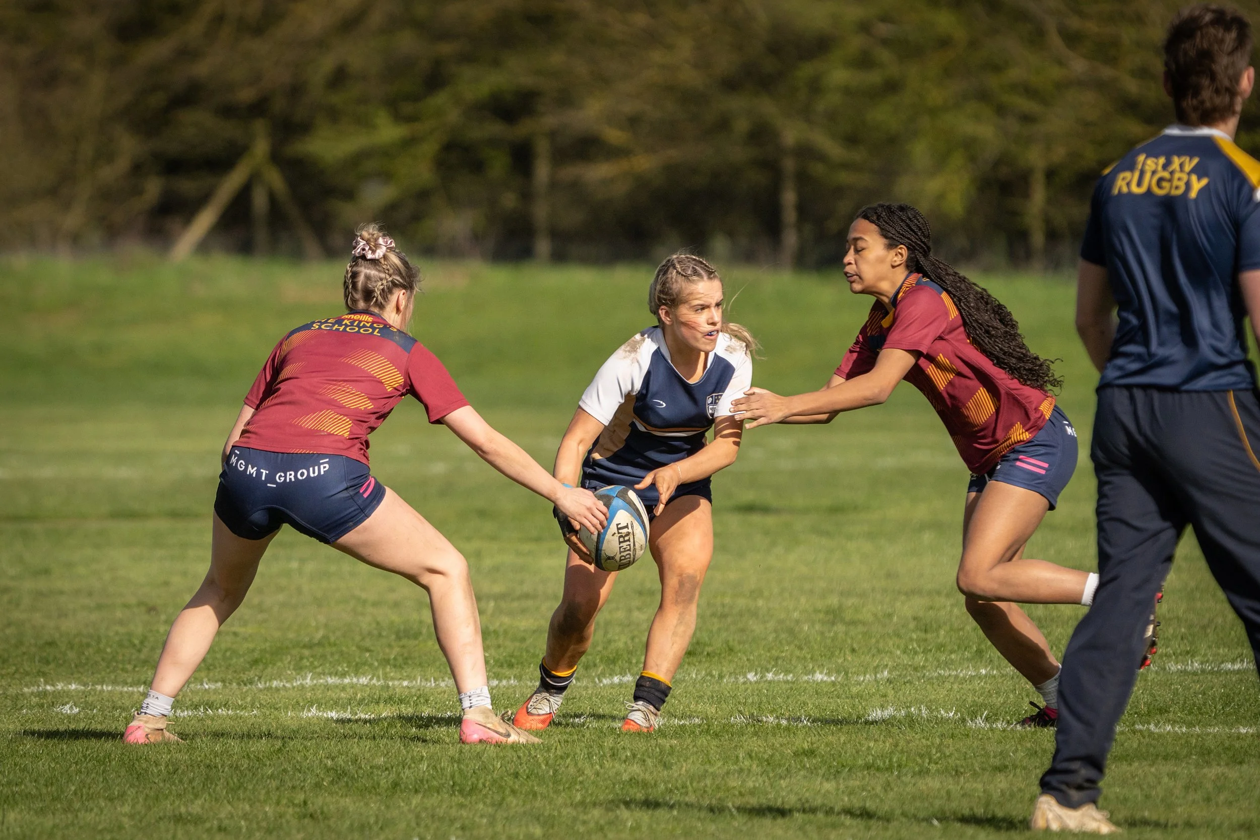 Women playing rugby on a grassy field with two women wearing maroon and navy jerseys and one woman in a navy jersey, with trees in the background.