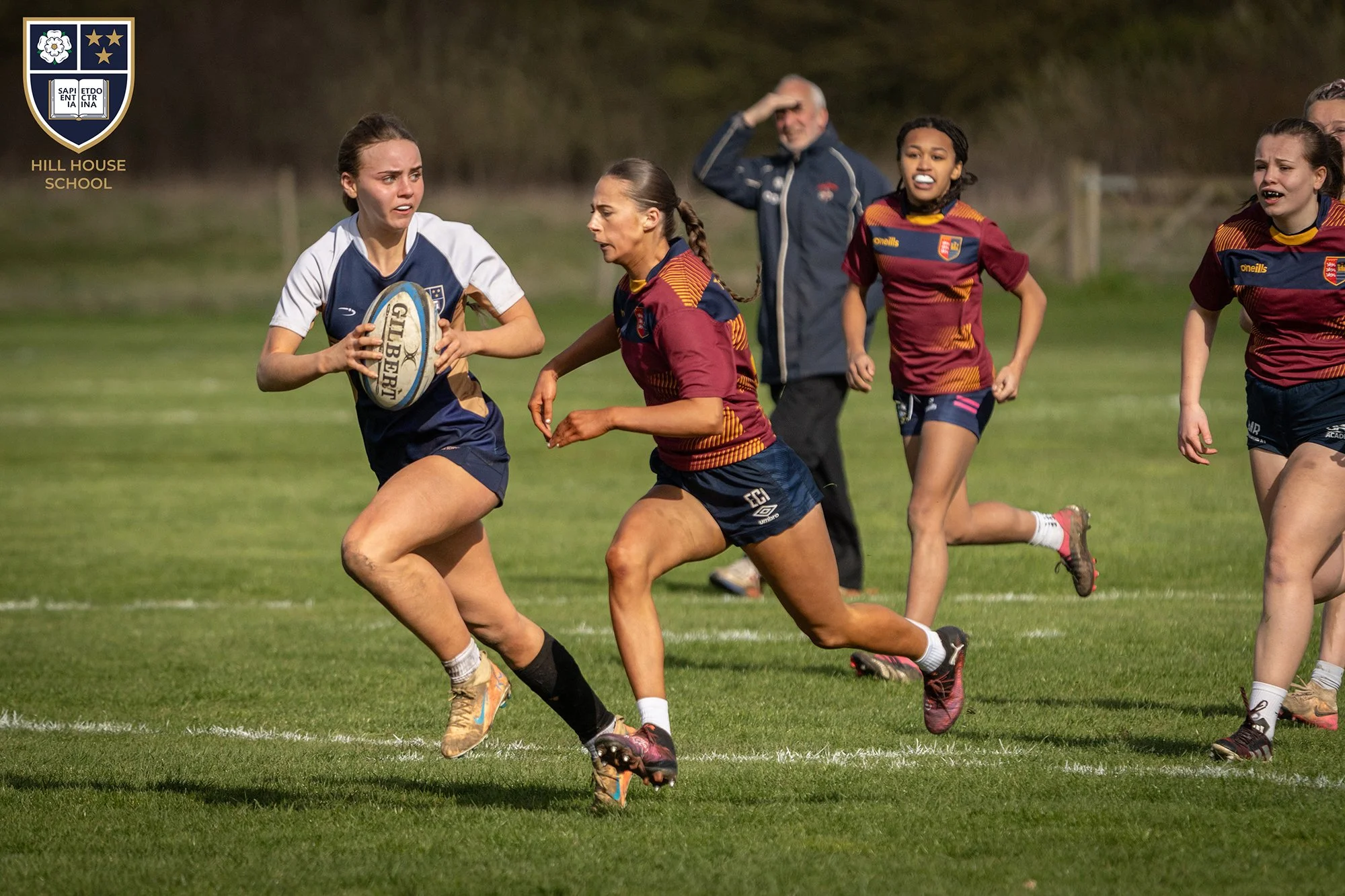 Women playing rugby on a grassy field with some players wearing maroon and navy jerseys and a girl holding a rugby ball. Two men are in the background, one saluting or shielding his eyes.