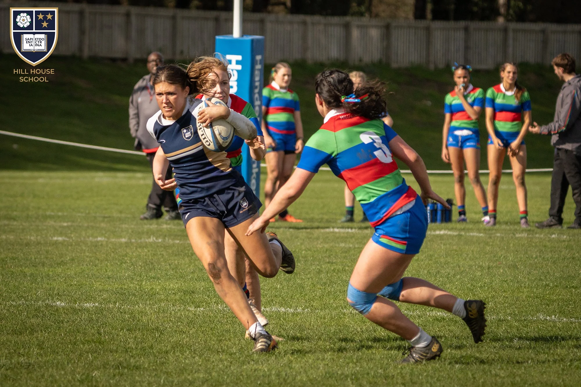Girls playing rugby on a grassy field, one girl in a navy blue and gold uniform with the ball, and another girl in a colorful striped uniform trying to tackle her. In the background, others watch, some in matching colorful uniforms, with a school emb