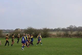 Children playing rugby on a grassy field with trees and overcast sky in the background.