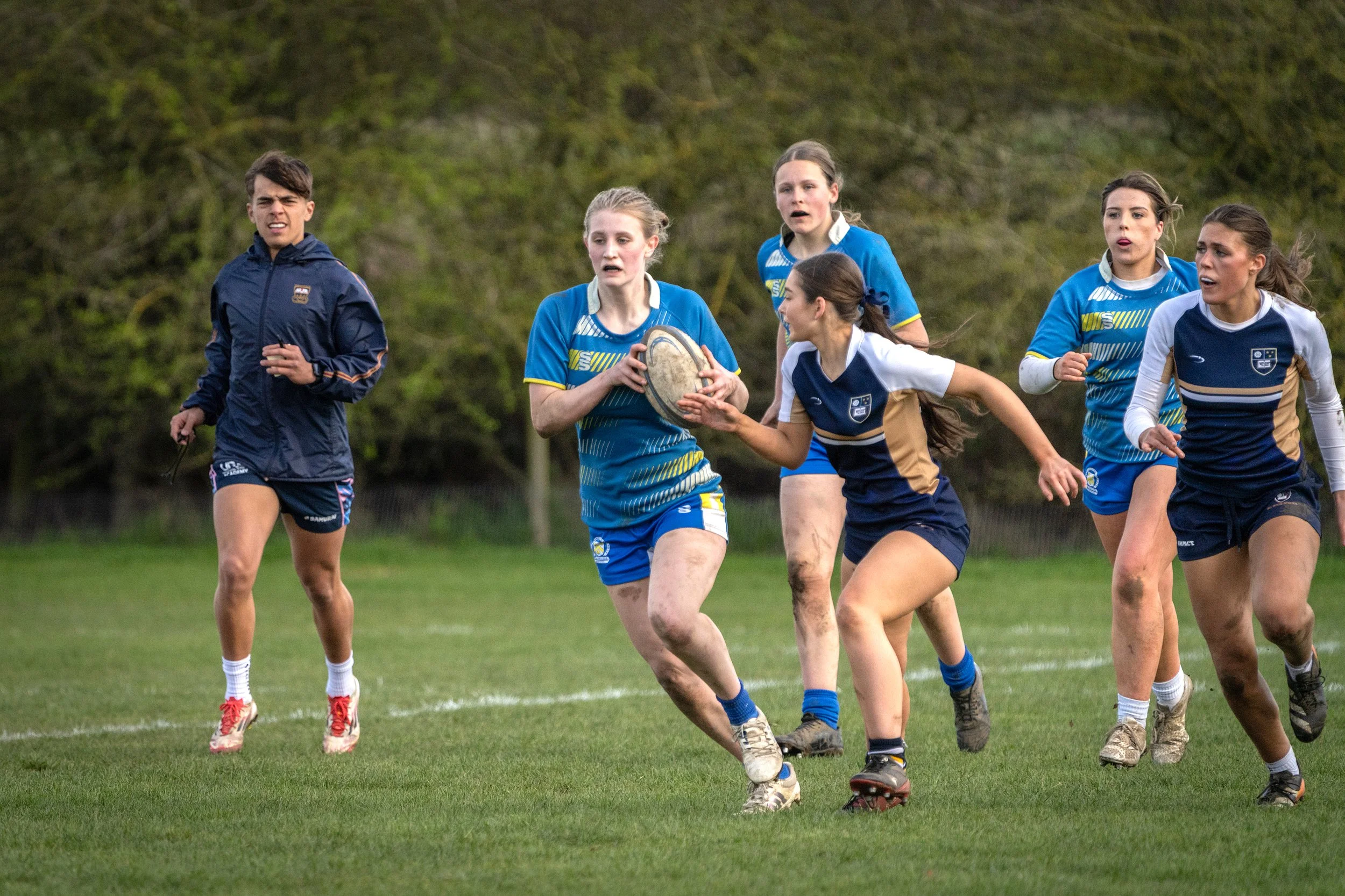 A group of young women playing rugby on a field, with one girl in blue holding the ball and others running alongside her.