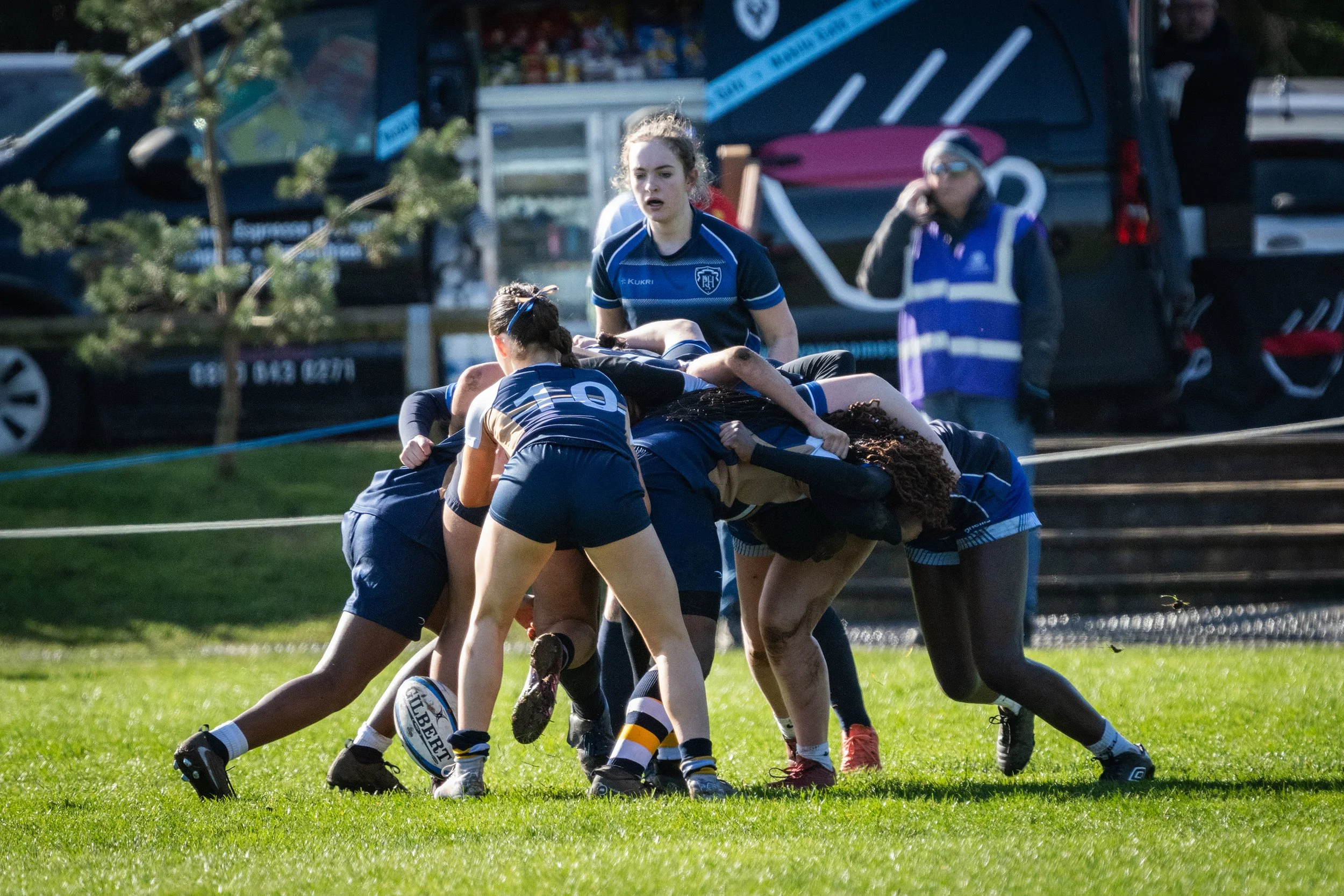 Rugby players engage in a scrum on a grassy field, with a referee observing nearby and cars in the background.