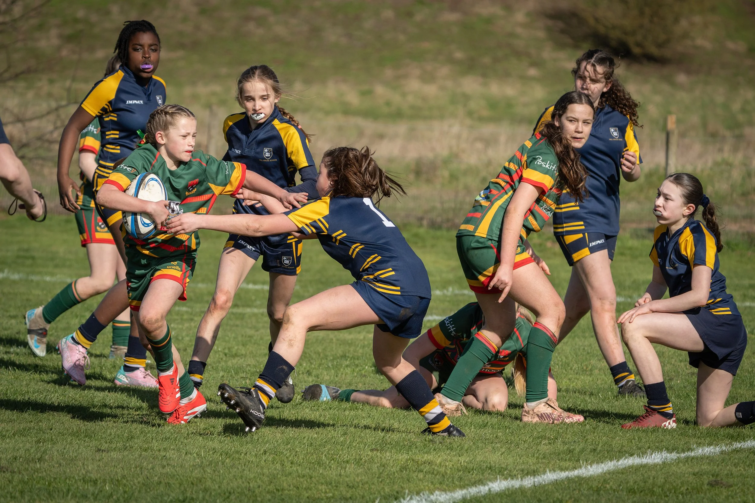 Young girls playing rugby on a grassy field, some wearing green and others blue and yellow uniforms, engaged in a tackle during a game.