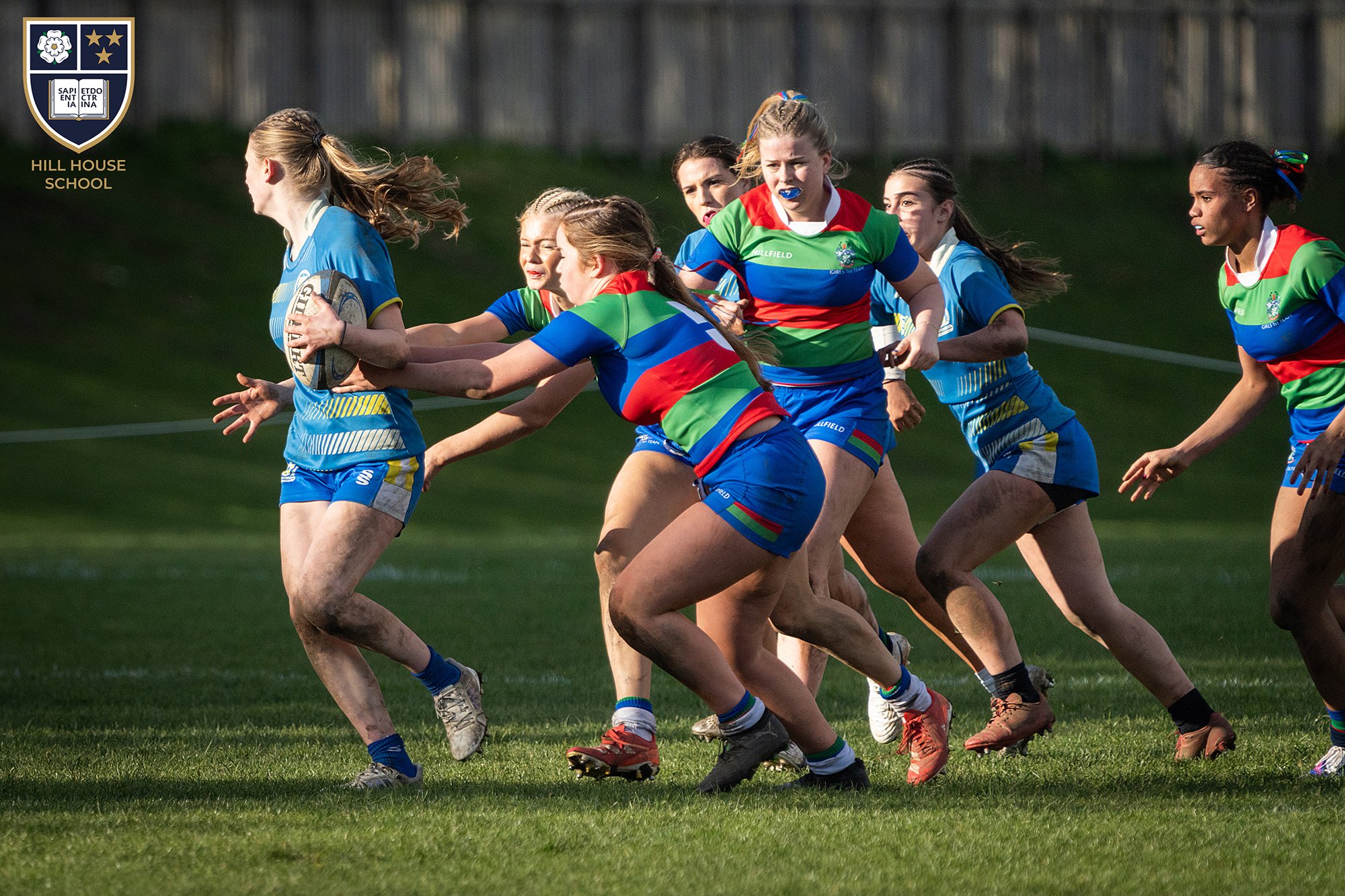 Girls playing rugby match on a grassy field, wearing colorful uniforms, with some muddy shoes and a wooden fence in the background.
