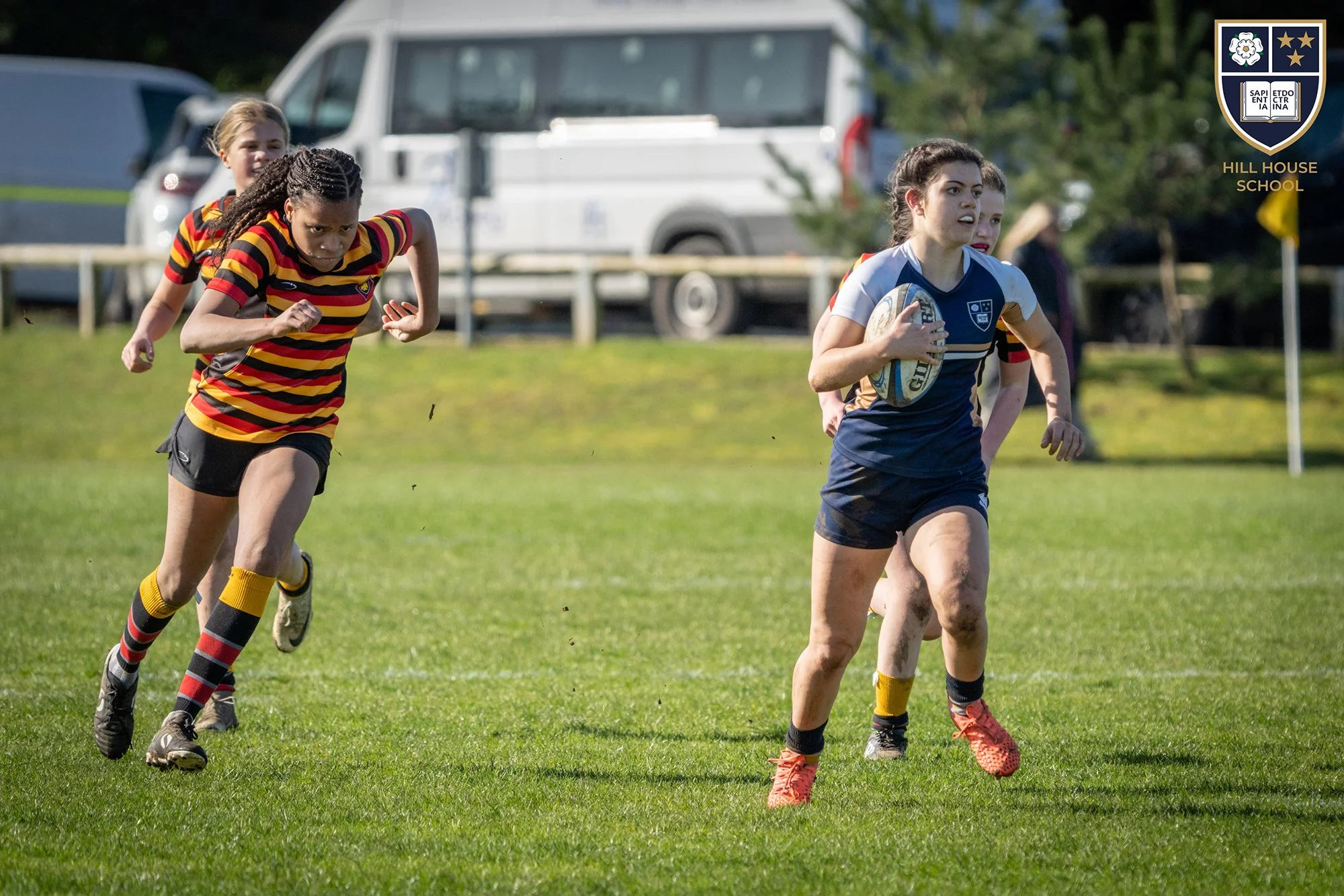 A group of young women playing rugby on a grassy field. One girl in a blue and white uniform is holding a rugby ball while running, and her teammates are following behind. The players are dressed in colorful sportswear. A vehicle and some trees are v