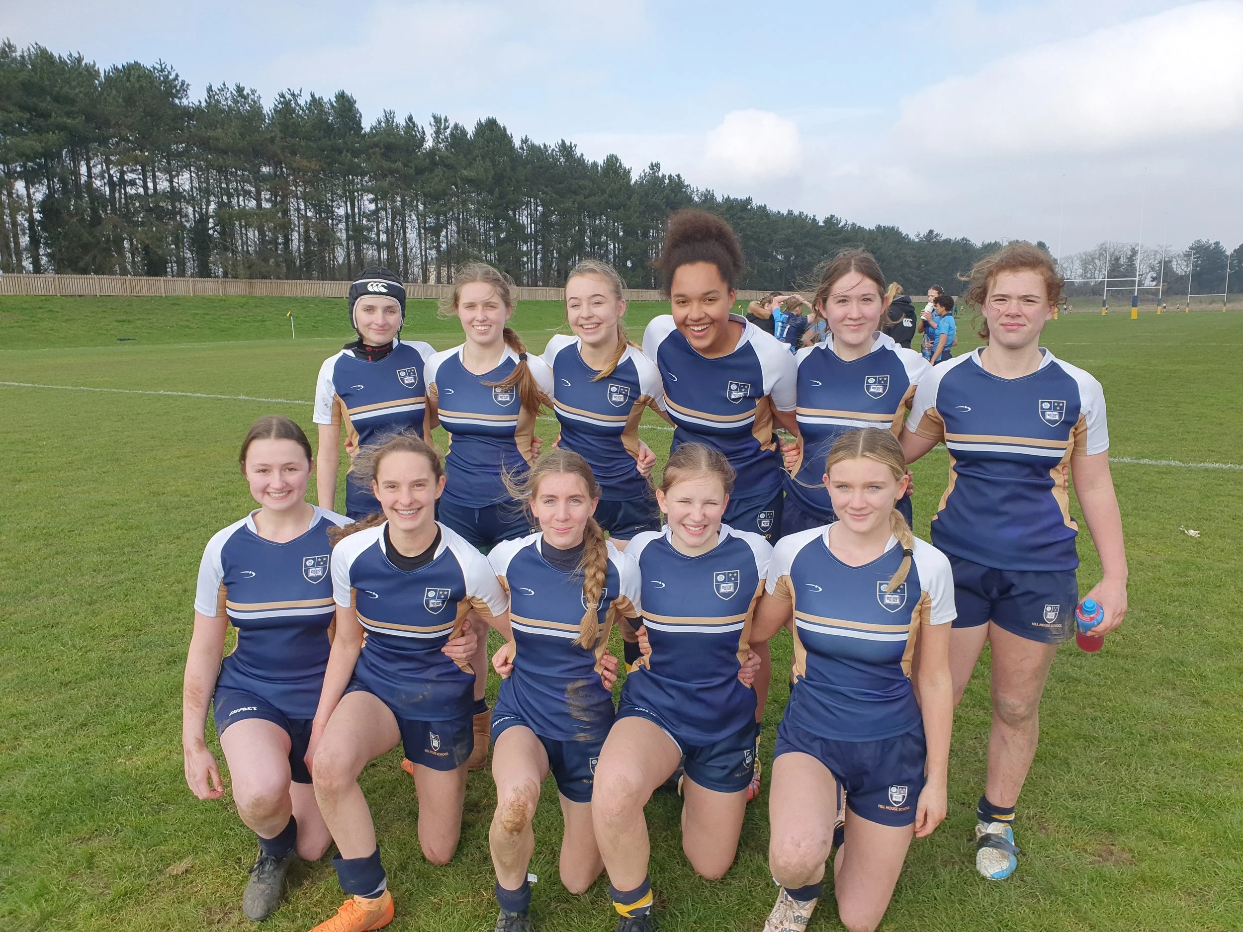 A group of young female rugby players in blue and white uniforms, standing on a grassy field with mountains and trees in the background.