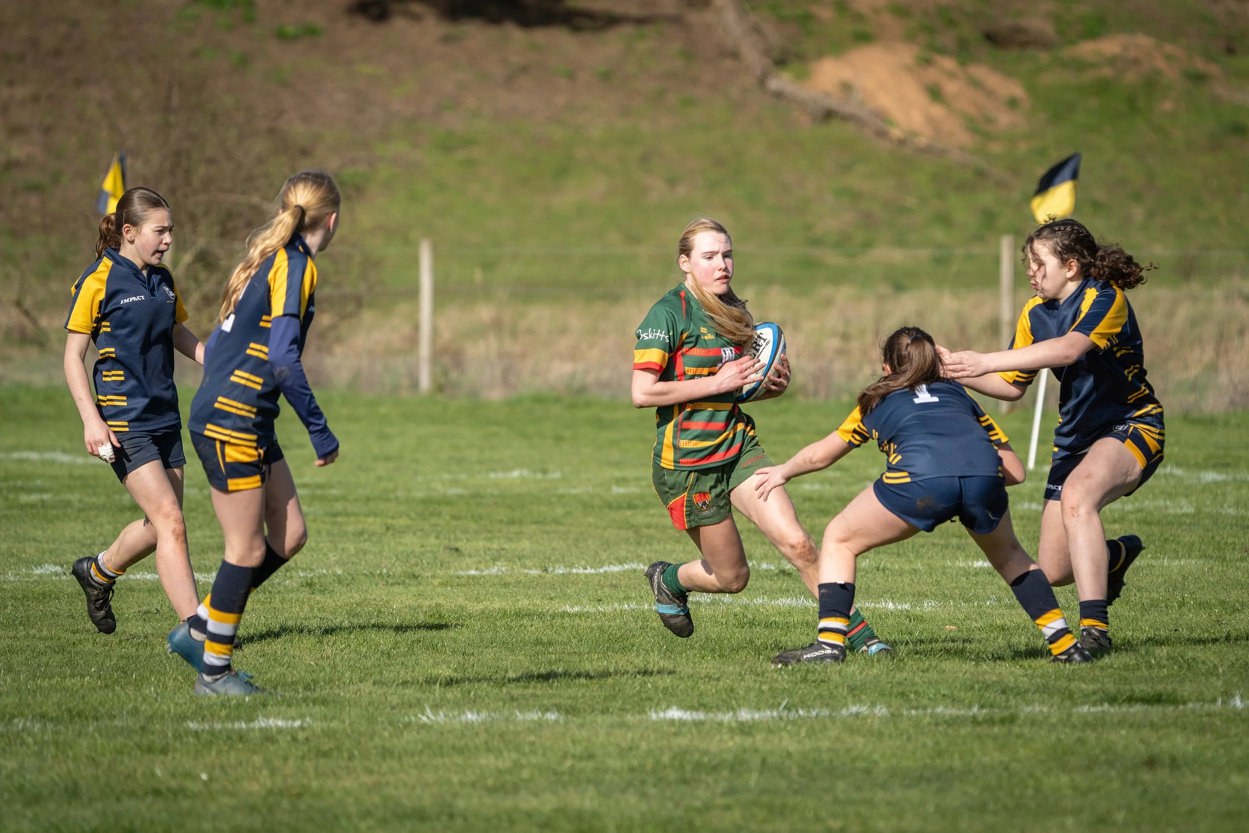A group of young women playing rugby on a grassy field, with one girl in a green and red jersey holding a rugby ball, surrounded by others in blue and yellow uniforms.