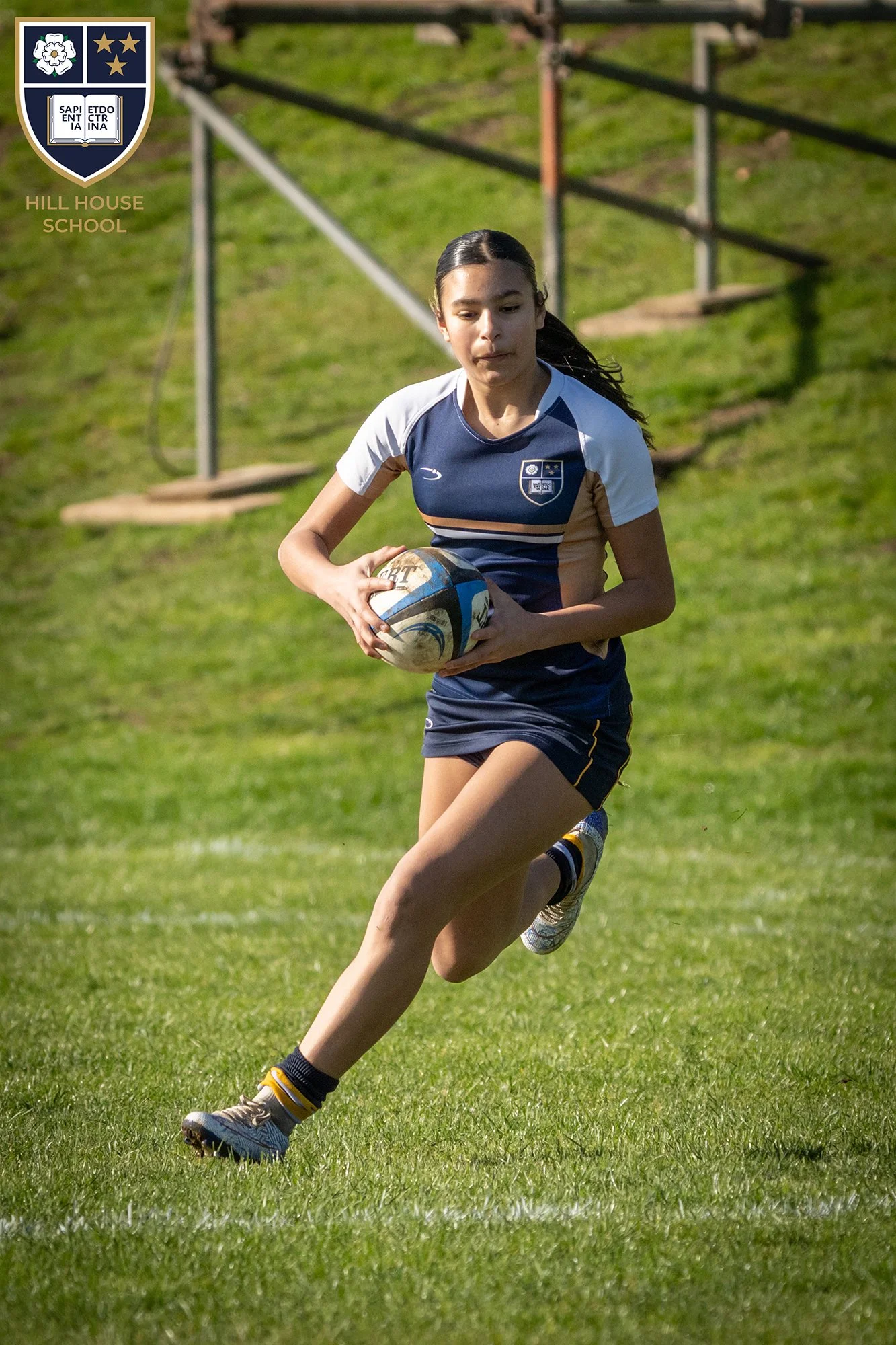 A young girl playing rugby on a grassy field, running with the ball tucked under her arm, wearing a blue and white sports uniform with a school crest, and running shoes.