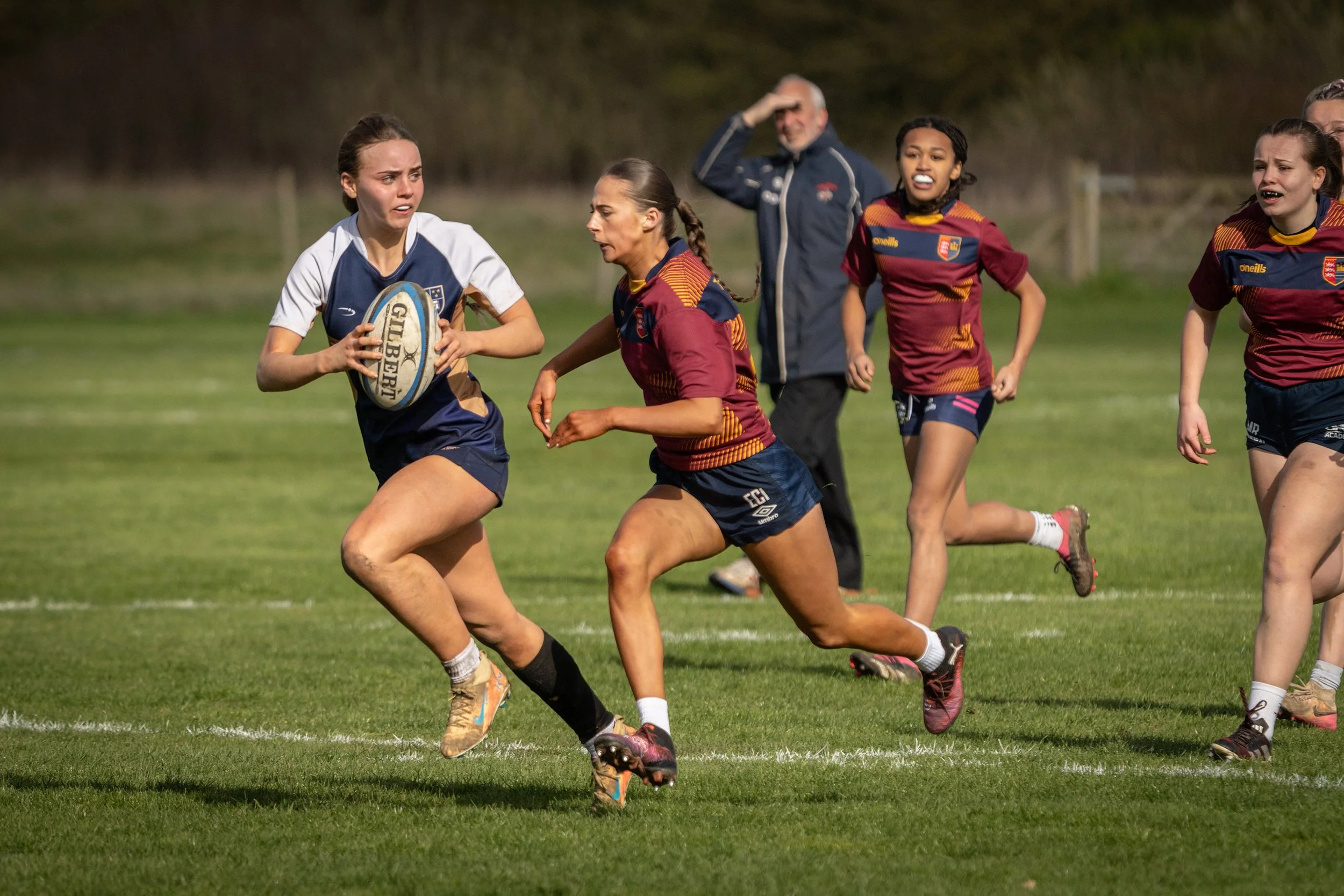 Women playing rugby on a grassy field, with some wearing maroon and navy jerseys and others in blue and white jerseys, and a man in the background.