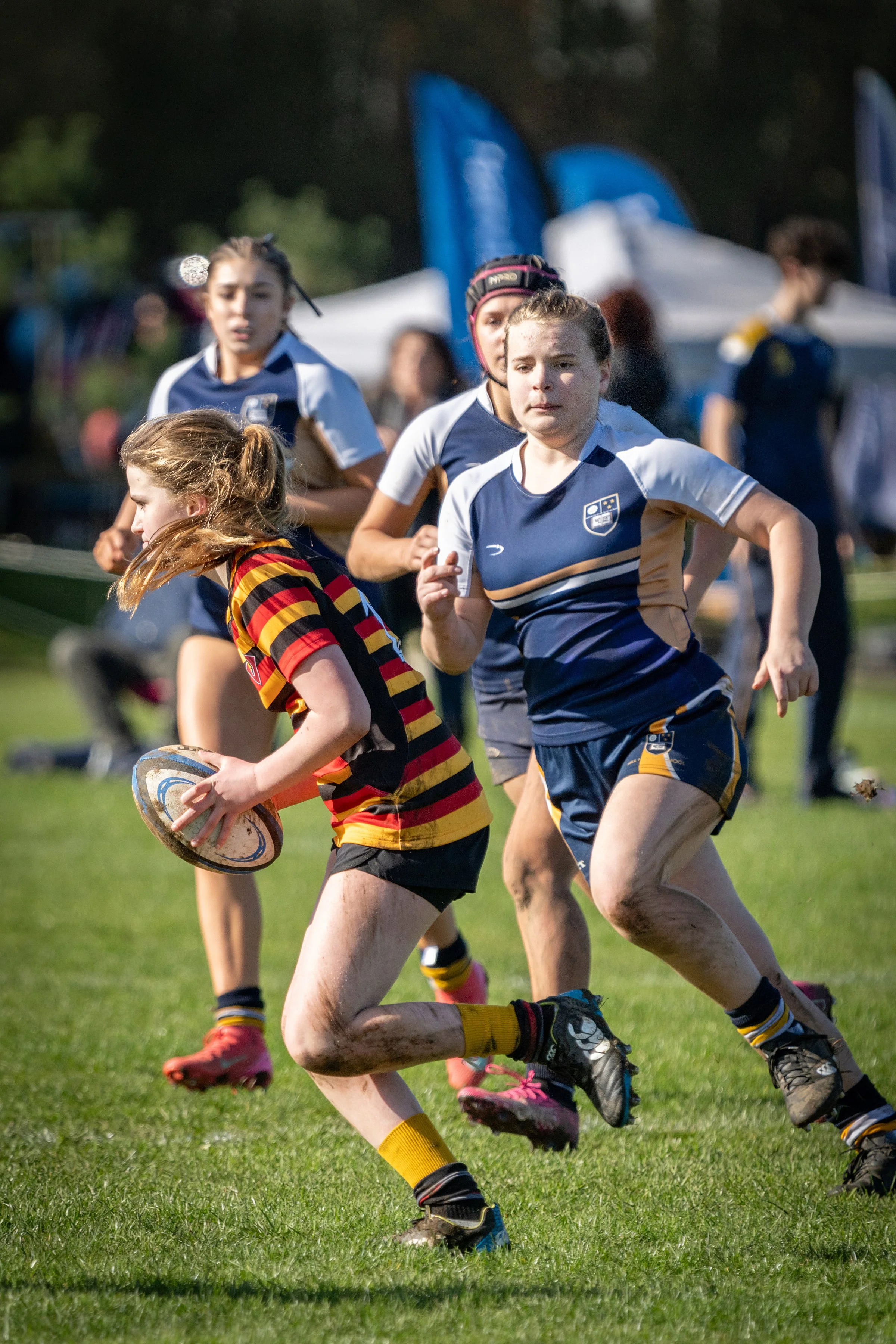 Girls playing rugby on a grassy field, some wearing team uniforms and one holding a rugby ball.