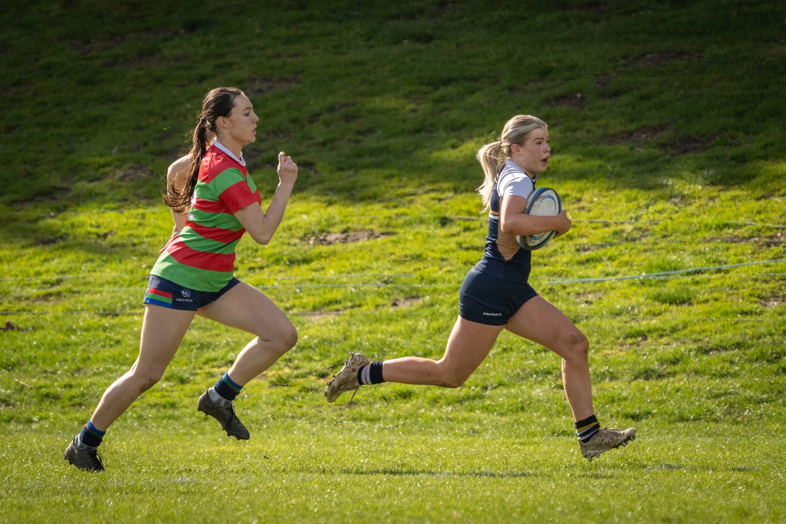 Two women running on a grassy field, one holding a rugby ball, with a hill in the background.