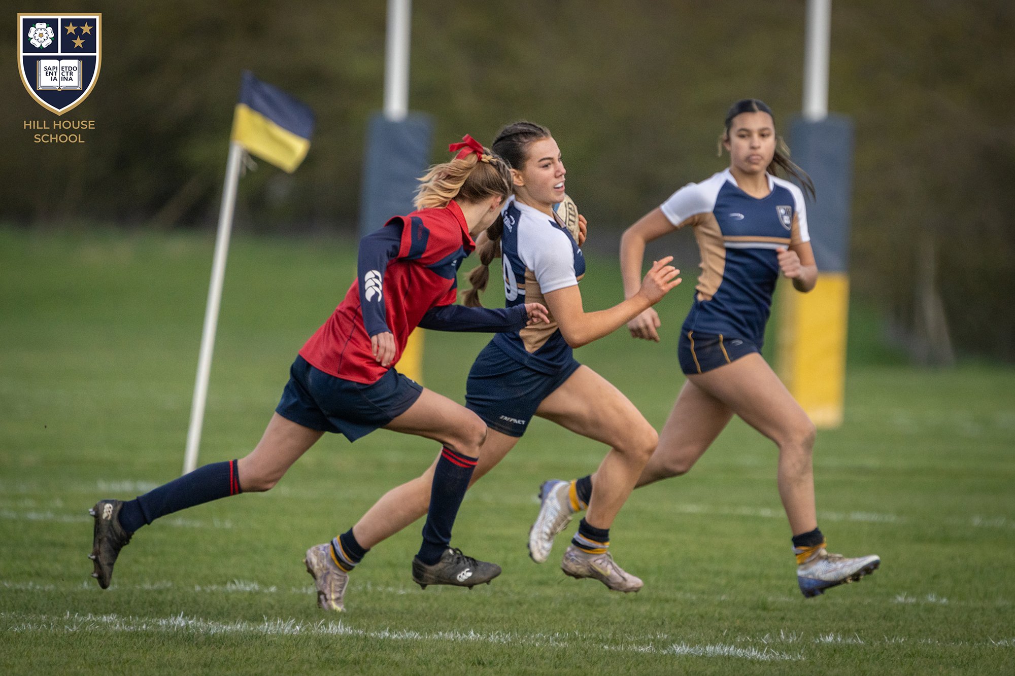 Three female rugby players running on a grassy field during a match, with a goalpost and a flag in the background, wearing team uniforms.