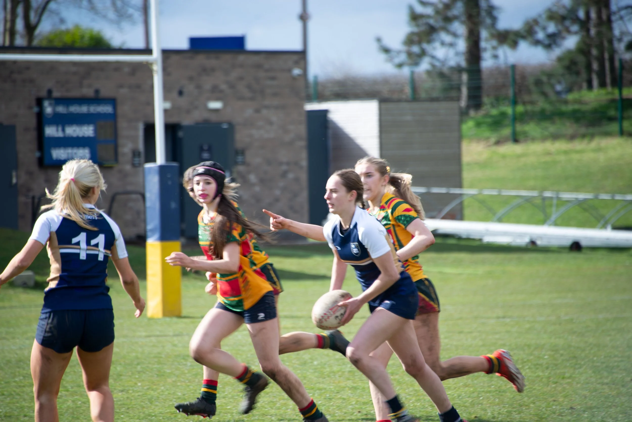 Rugby players in action on a grassy field with a brick building and trees in the background.