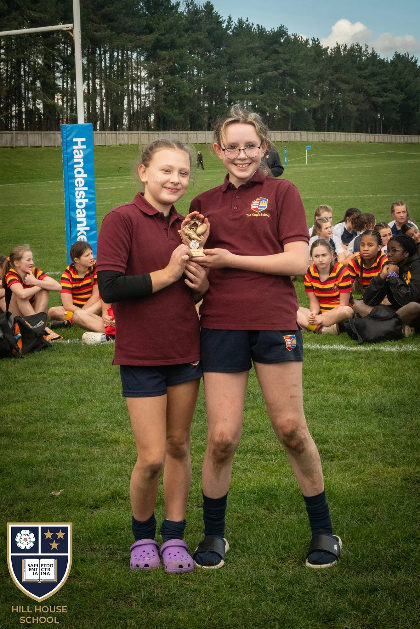 Two girls in maroon school shirts holding a small trophy on a sports field, with other students sitting in the background.