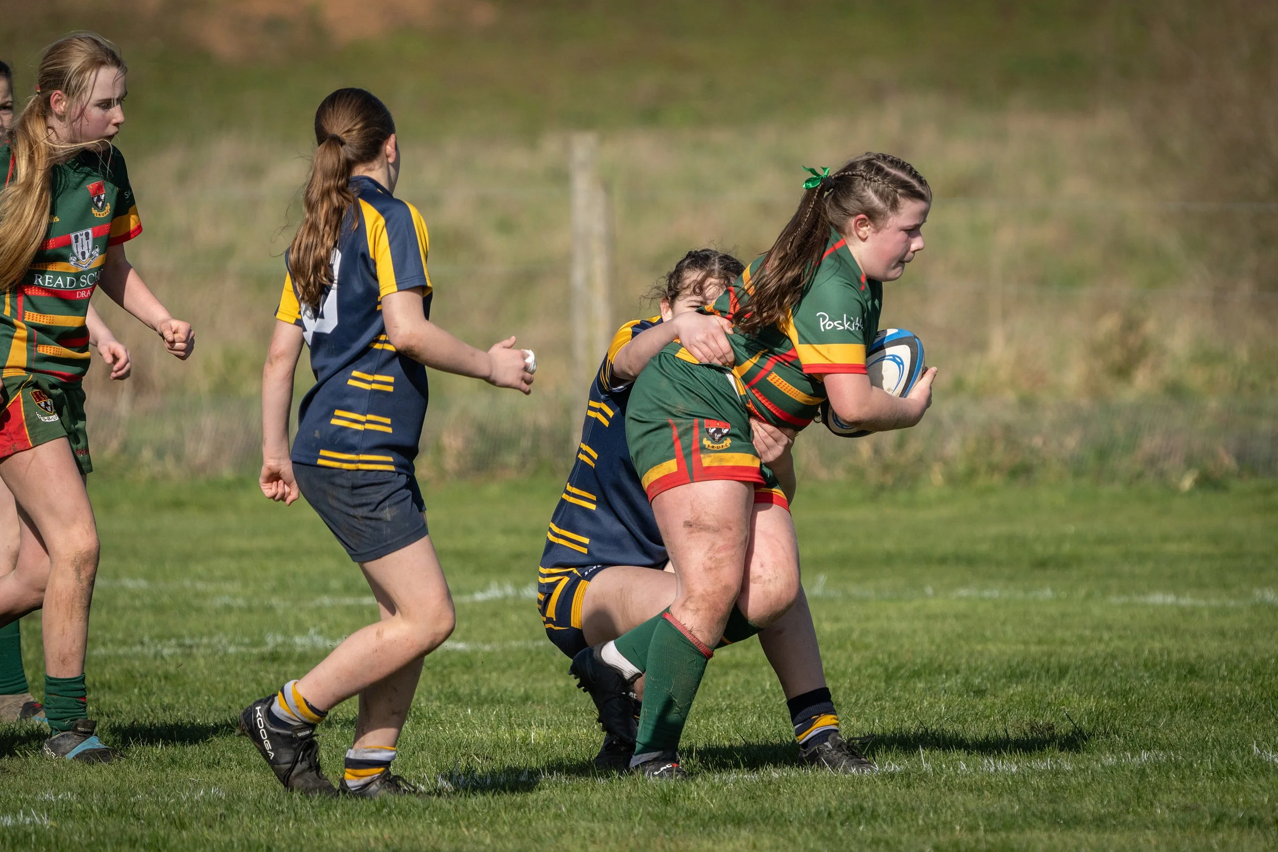 Girls playing rugby on a grassy field, with one girl in green holding a rugby ball while others wearing blue and yellow jerseys chase her.