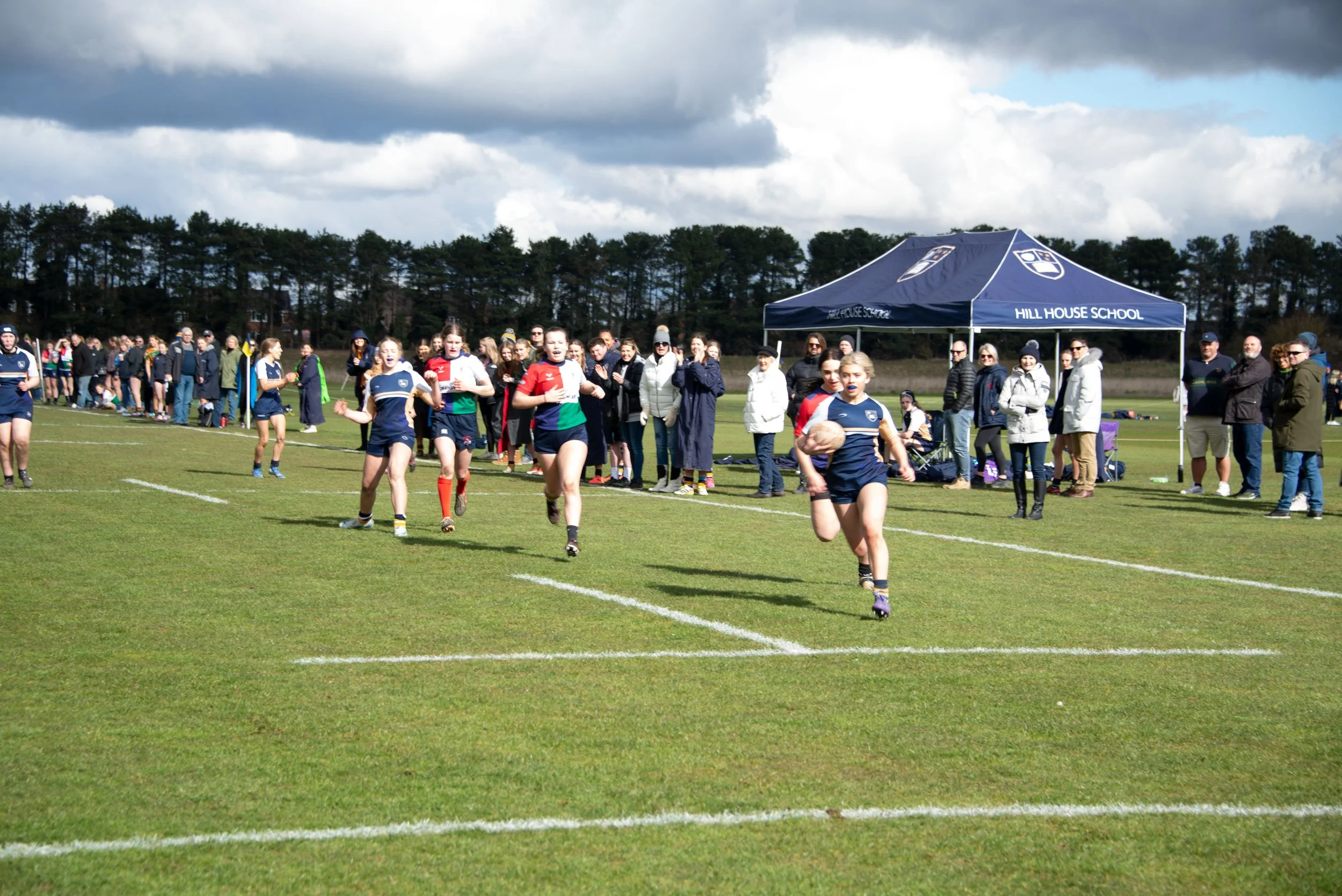 Young girls playing rugby on a grassy field during a school sports event, with spectators and a tent labeled 'Hill House School' in the background under a partly cloudy sky.