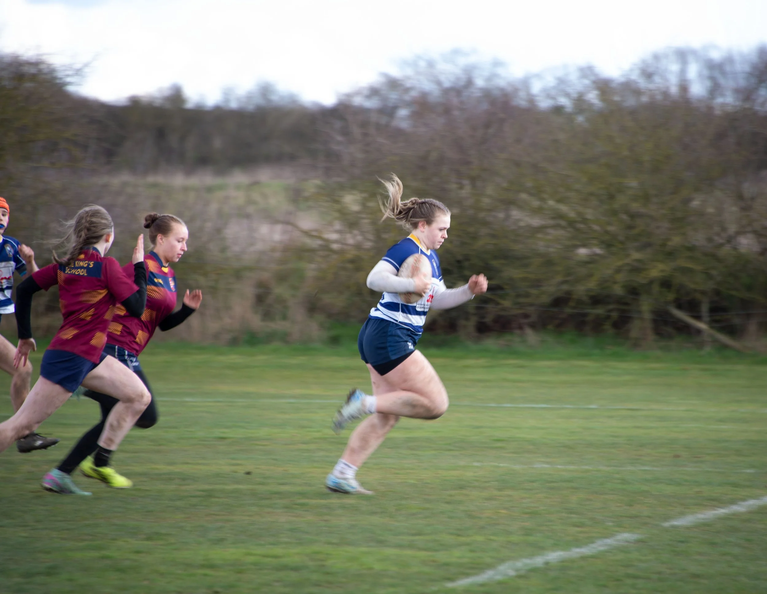 A group of young girls playing rugby outdoors on a grassy field, with trees in the background.