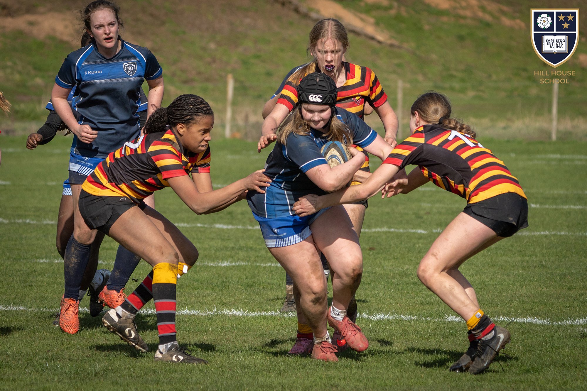 Girls rugby players in action during a match, wearing colorful team uniforms. The scene is on a grassy field with some hills in the background, and one girl in the center is holding a rugby ball.