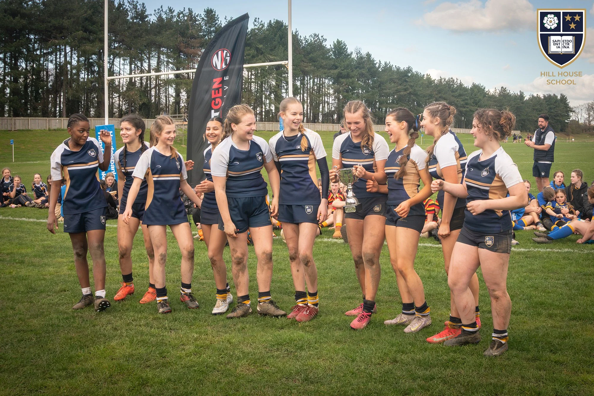 Young female rugby players celebrating with a trophy on a grassy field, with team members seated behind them, some muddy, and a coach or staff member standing on the right. There are flags and trees in the background, with Hill House School logo in t