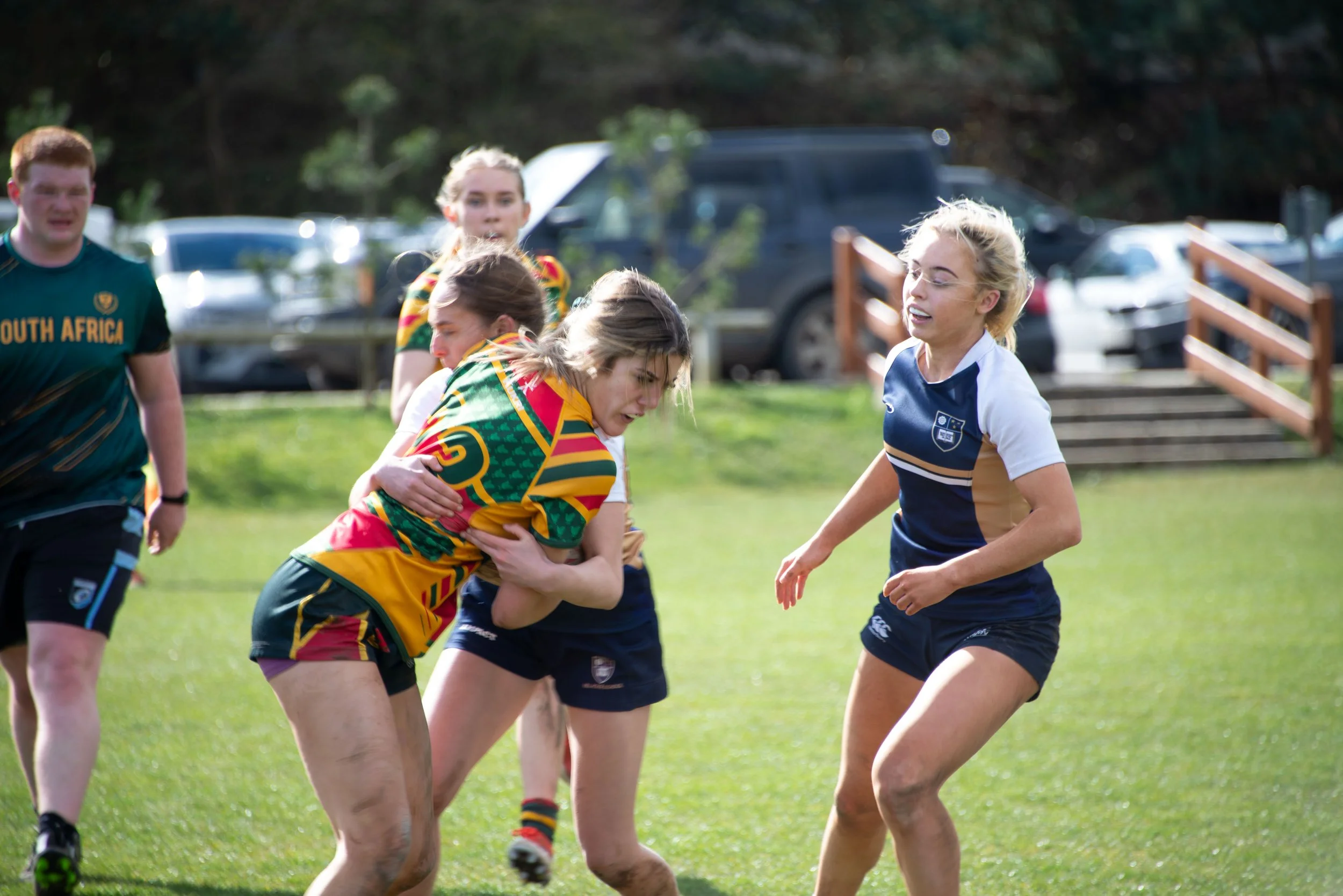 Women playing a rugby match outdoors, wearing jerseys, one in colorful pattern and others in navy blue, with one woman holding her stomach, indicating a physical contest.