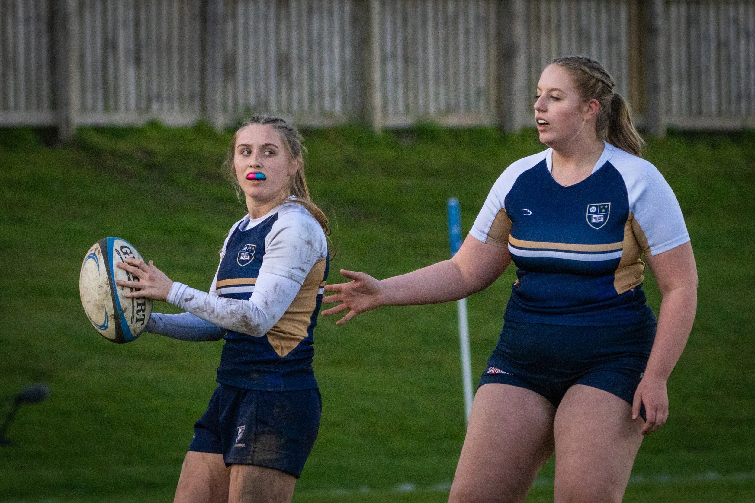 Two female rugby players in blue and gold jerseys practicing on a grassy field during daytime. One is holding a rugby ball, while the other is standing nearby. They are near a fence in the background.
