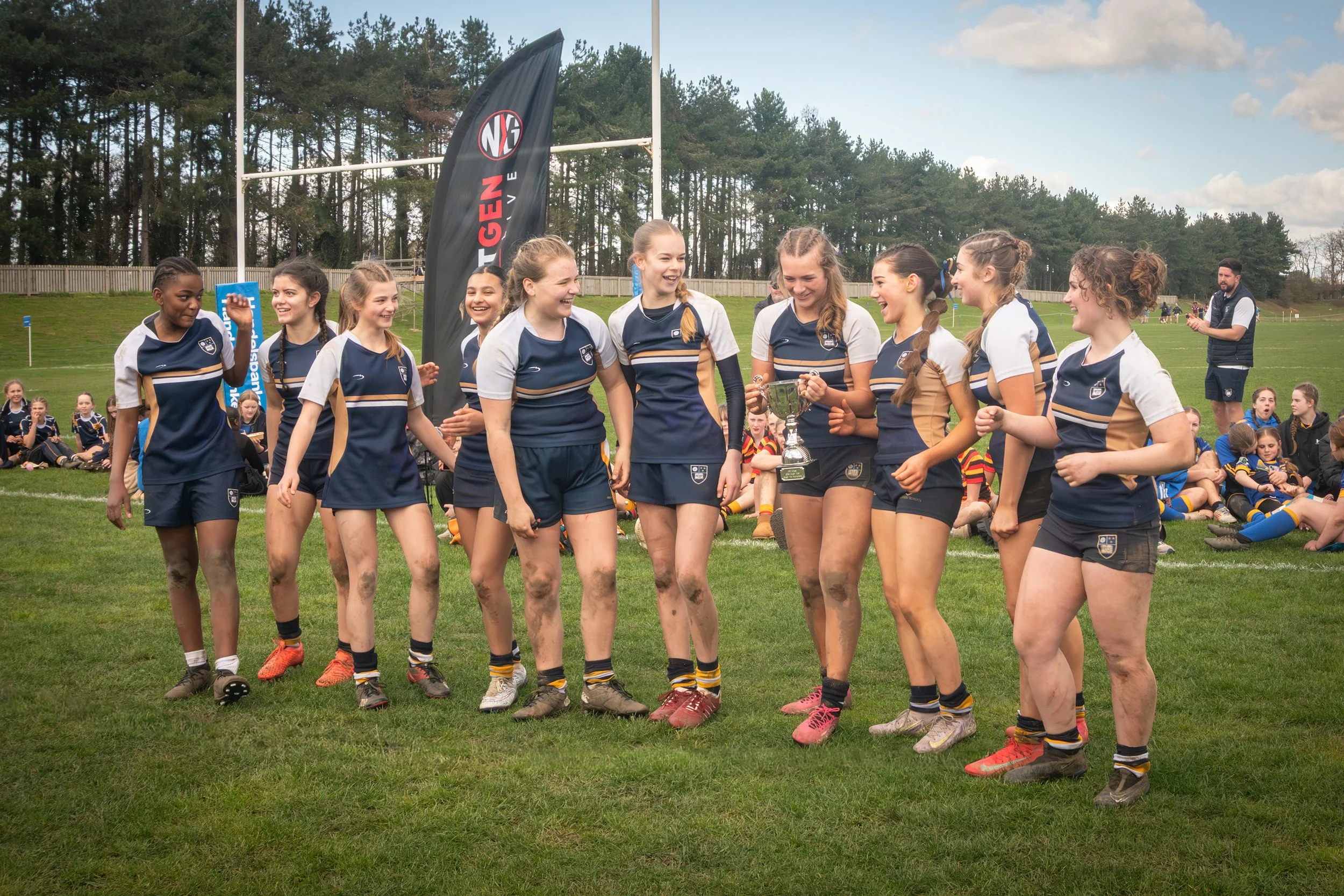 A group of young girls in sports uniforms are celebrating on a soccer field, with some holding a trophy. They appear happy and are smiling, with muddy legs and soccer shoes. In the background, there are more players and coaches, a fenced field, and t