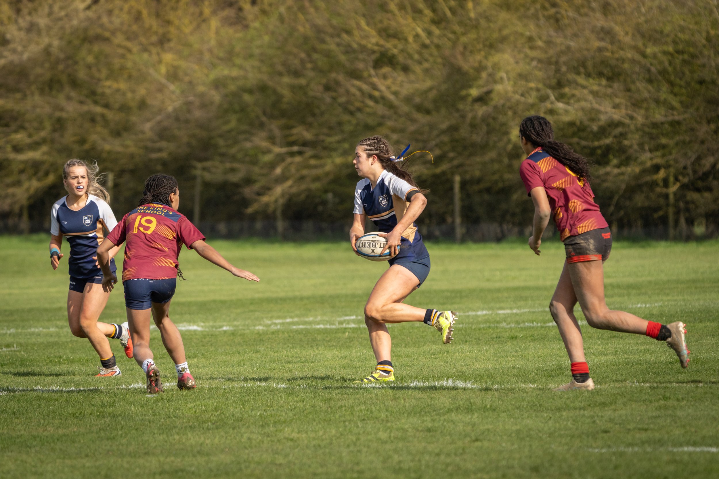 A women's rugby game on a grassy field with players in blue and maroon jerseys, and one player holding a rugby ball.