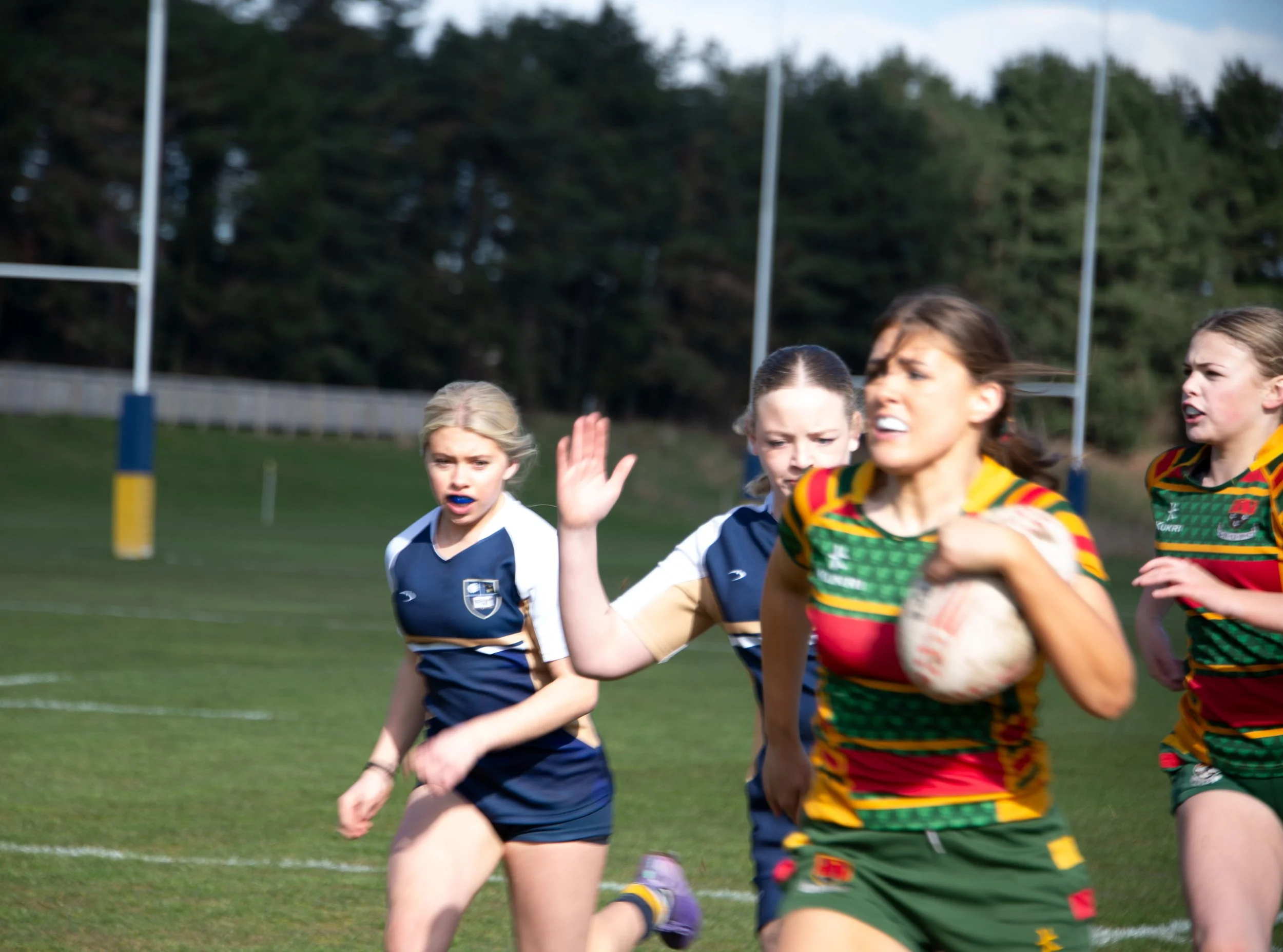 Women and children playing rugby on a grassy field with goal posts and trees in the background.