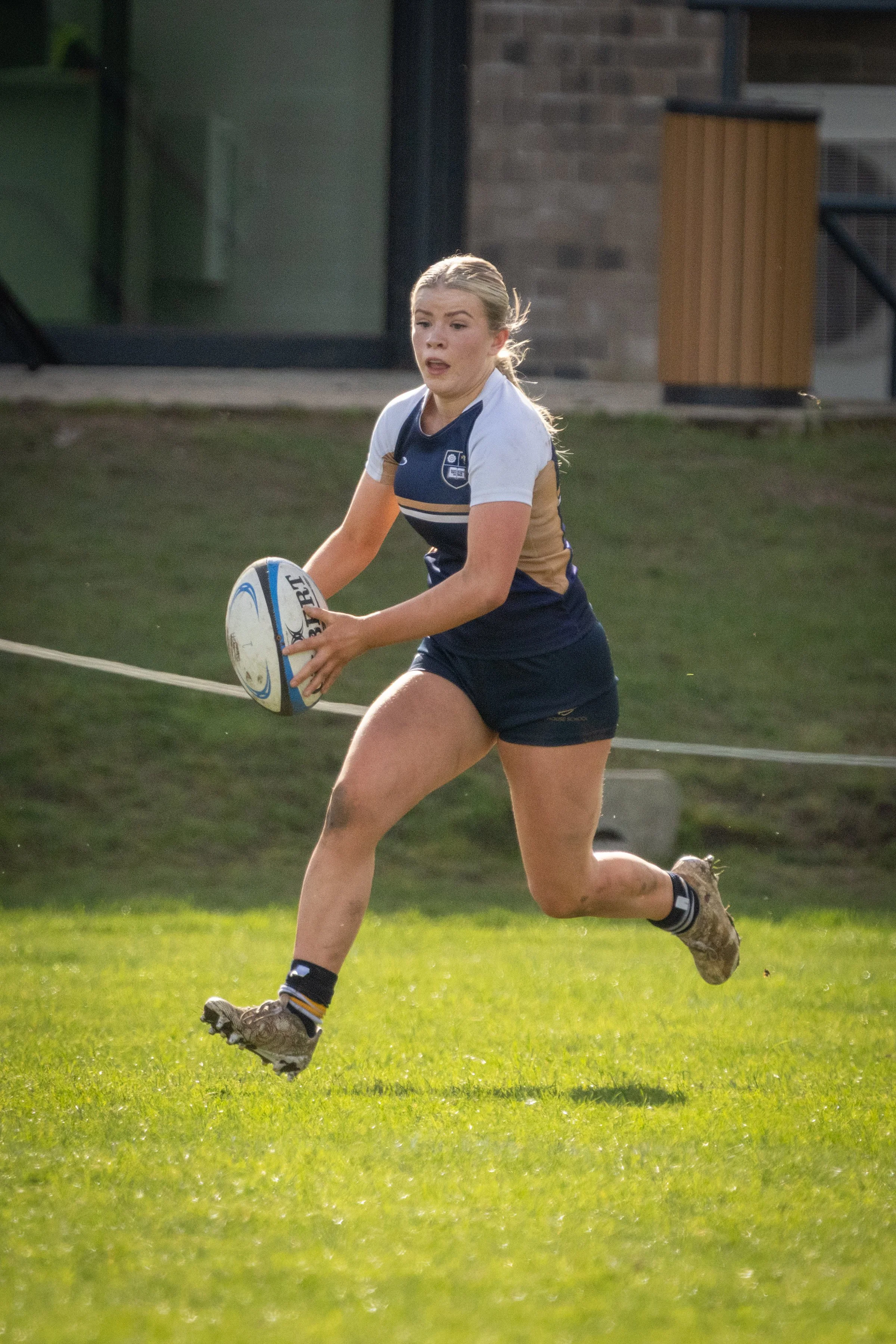 A female rugby player running on a grassy field while holding a rugby ball.