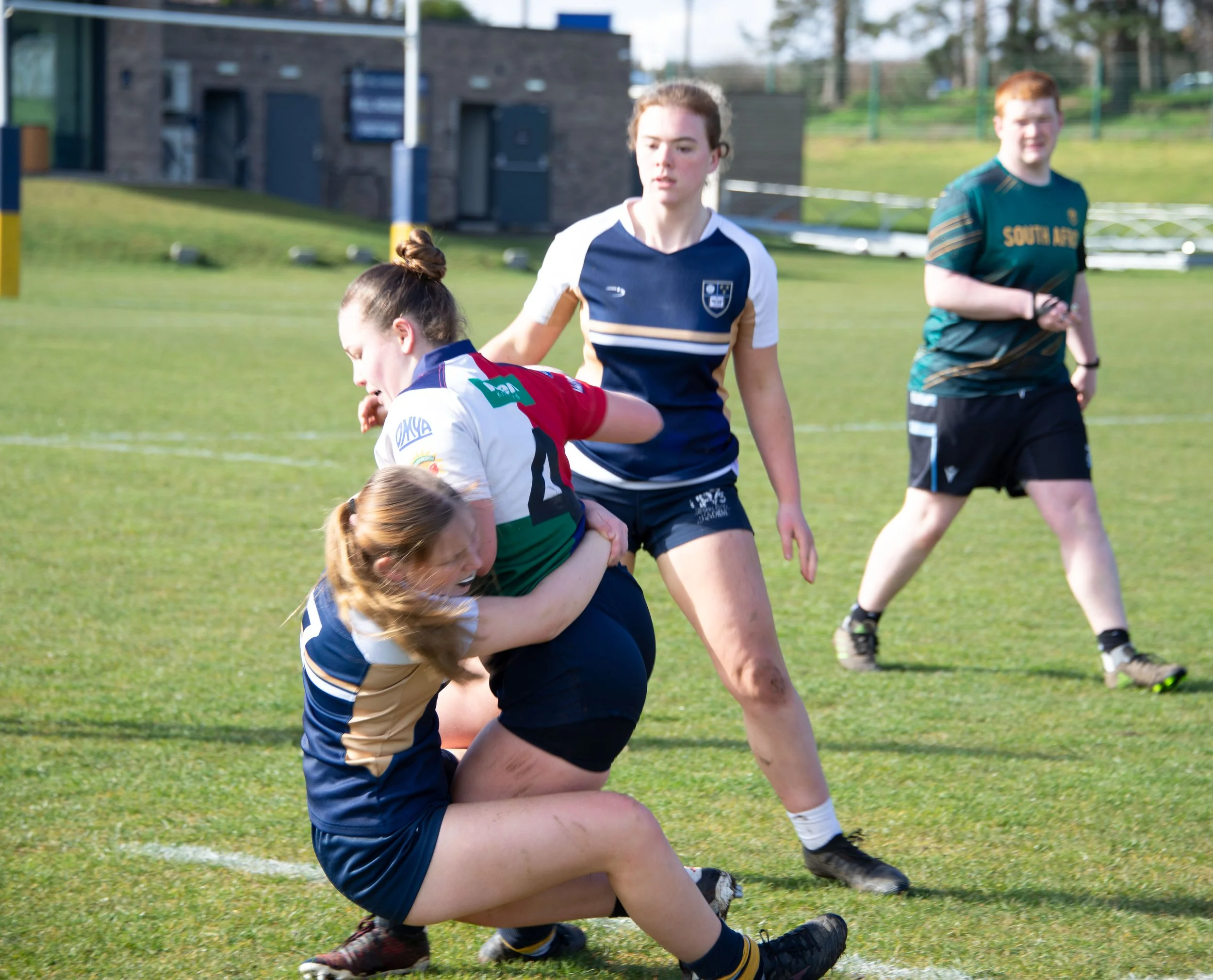 Rugby players on the field, one in a white and red jersey, others in dark jerseys, with a grassy field and building in the background.
