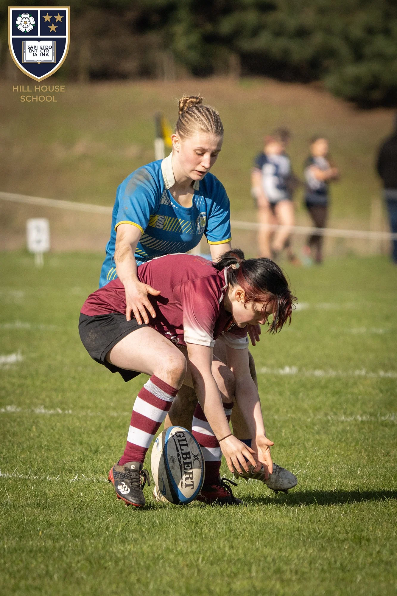 A rugby game with players on the field. One player in a maroon and white uniform is on her hands and knees with a rugby ball nearby. A player in a blue uniform is behind her, appearing to assist or cheer. The background shows a grassy field, some blu