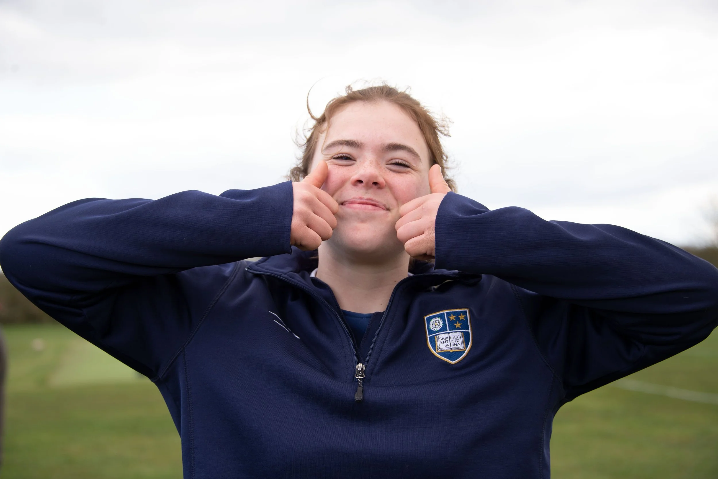 A smiling young girl with curly red hair giving two thumbs up outdoors on a cloudy day, wearing a dark blue sports jacket with a sports team emblem on the chest.