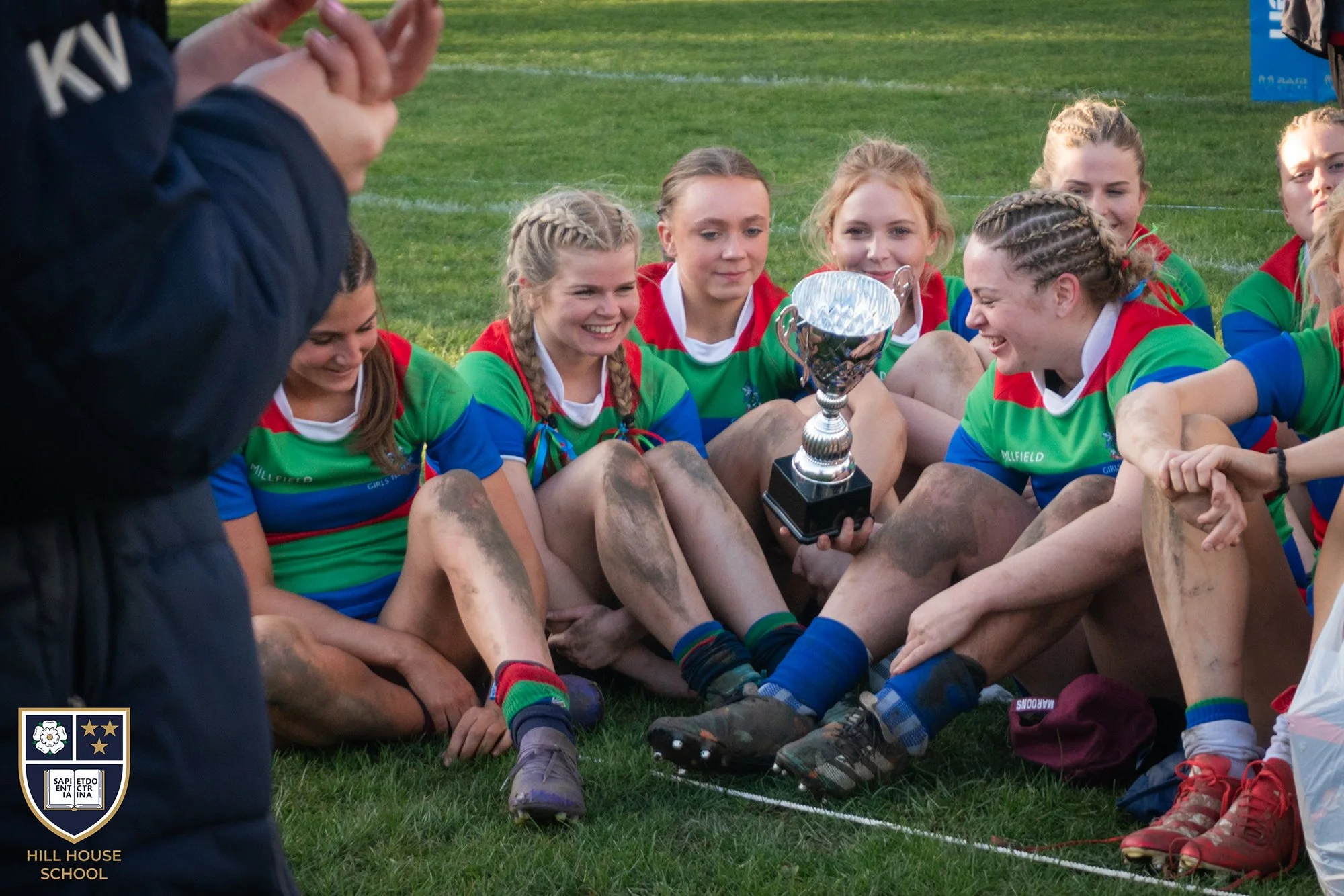 A group of young women in colorful sports uniforms sitting on a grassy field, holding a shiny silver trophy, smiling and celebrating a victory in a sports event.