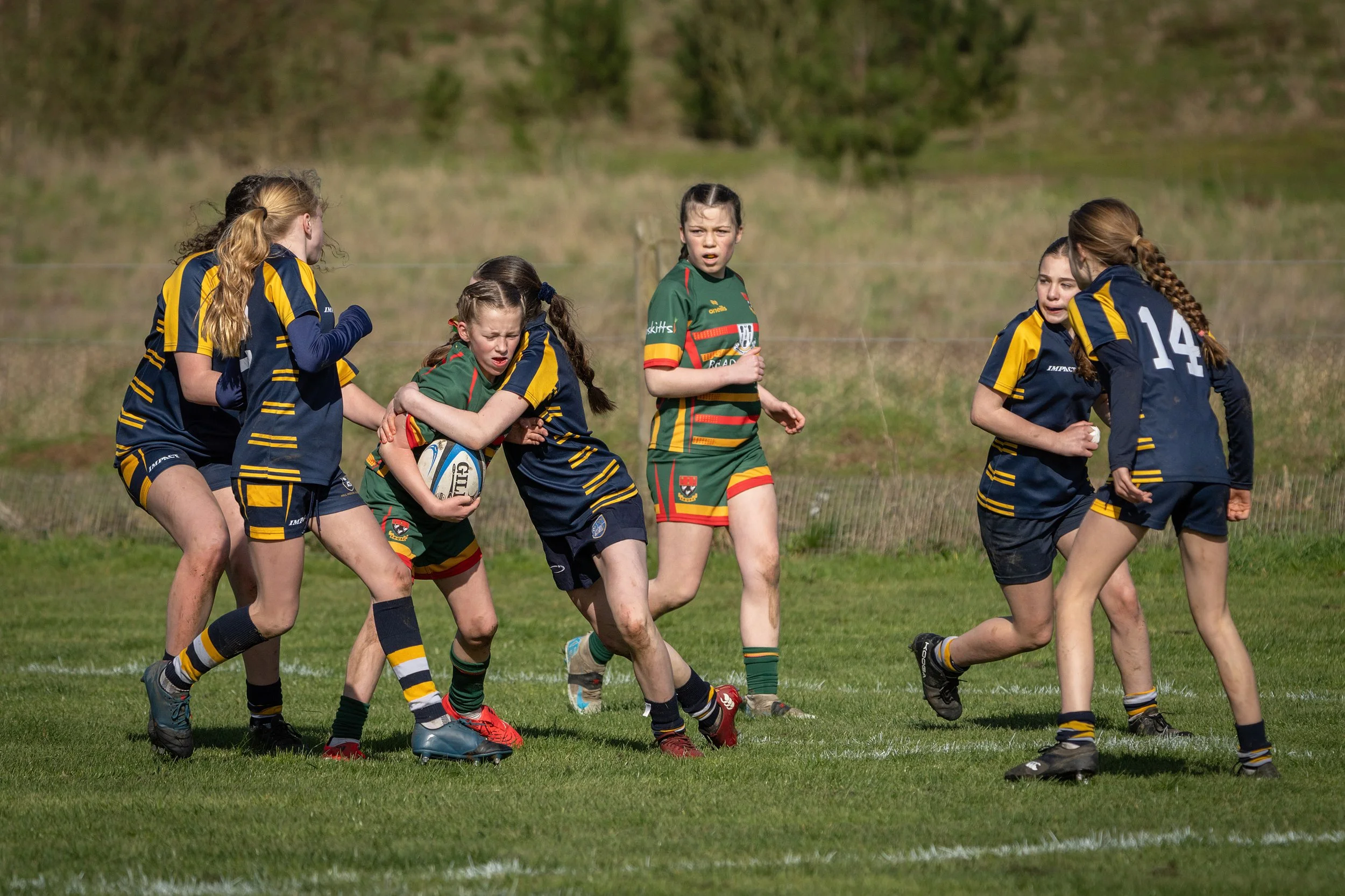 Girls playing rugby on a grassy field with a fence and trees in the background.