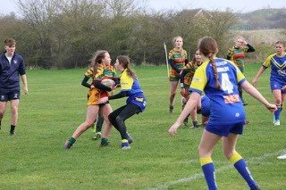 Women playing rugby on a grassy field with children and a few spectators in the background.