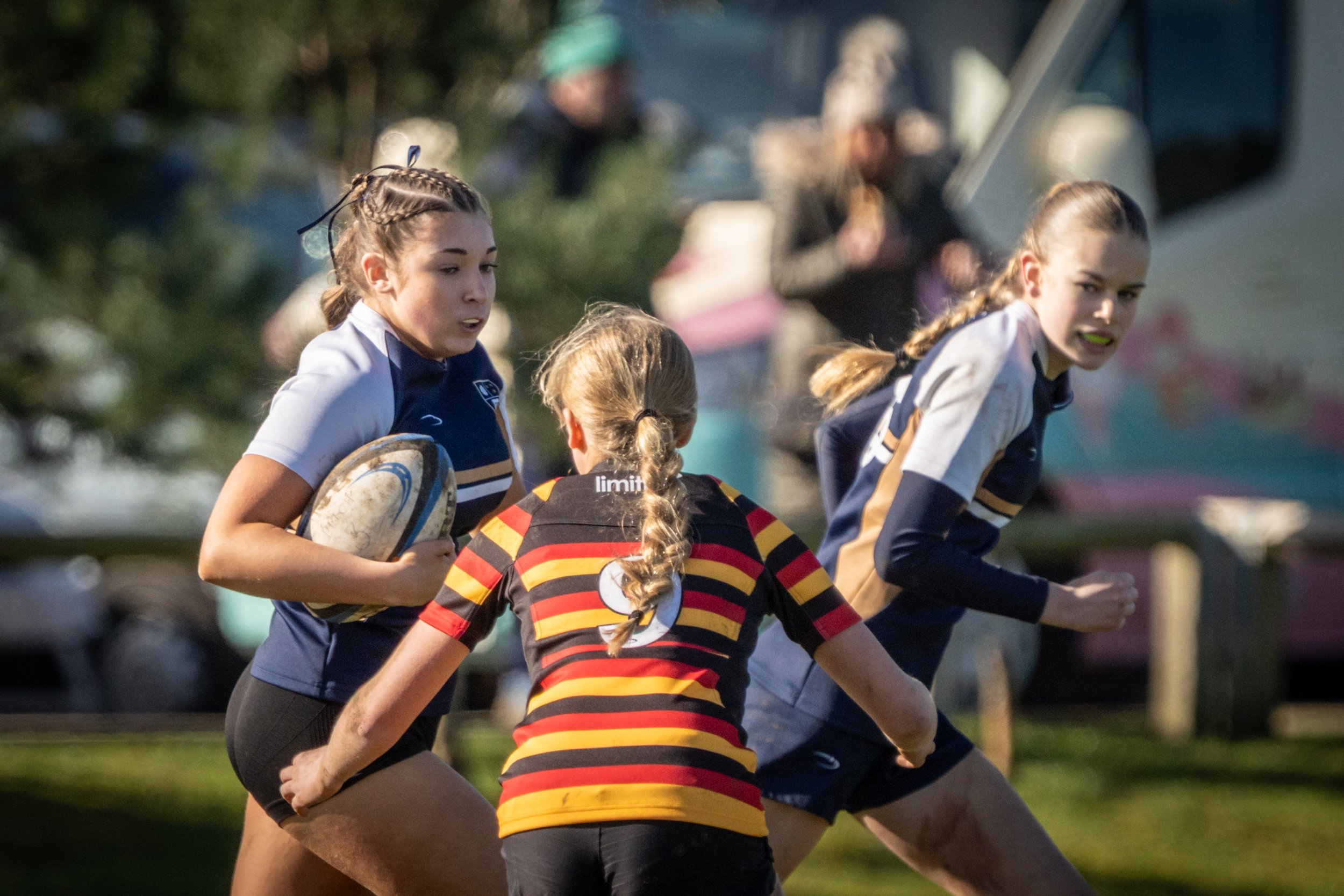Women and girls playing rugby outdoors during daytime, some wearing striped jerseys and athletic clothing, engaged in a rough game.