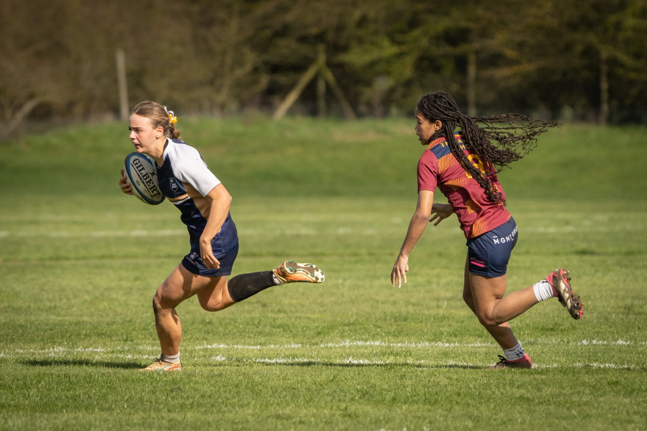 Two female rugby players in action on a grassy field, one holding a rugby ball and running, the other chasing, with trees in the background.