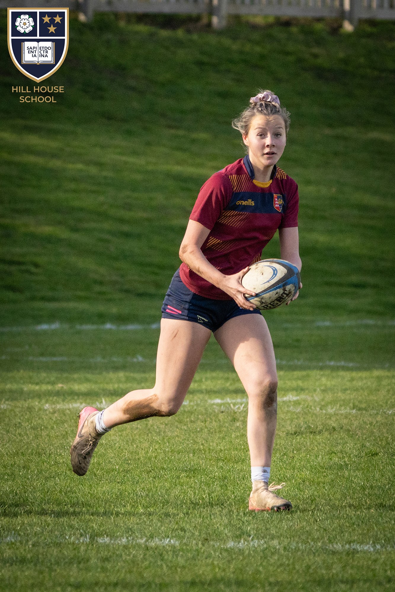 A female rugby player with blonde hair tied up and a scrunchie, wearing a maroon and blue jersey, running on a grassy field while holding a rugby ball, with a logo in the top left corner of the image.