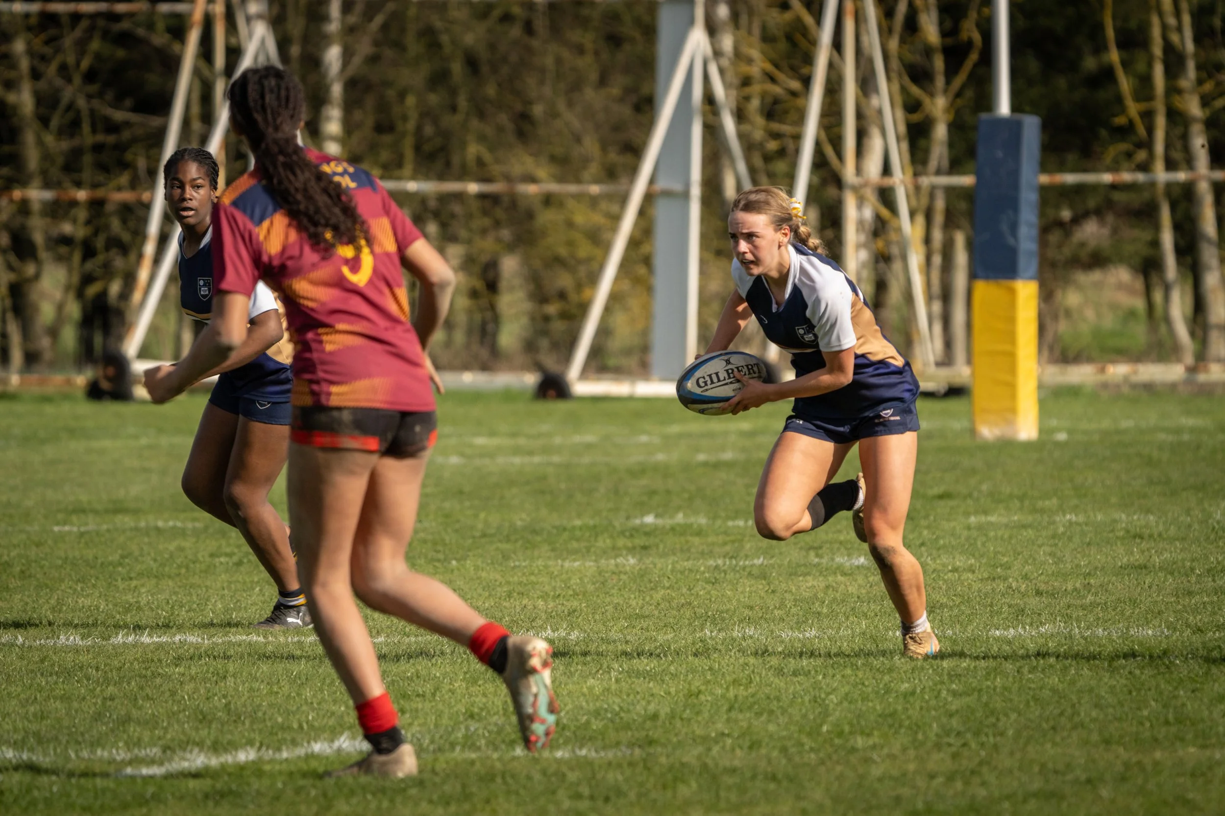 Rugby players in action during a game, with a girl holding the ball ready to run, on a grass field with goal post in the background.