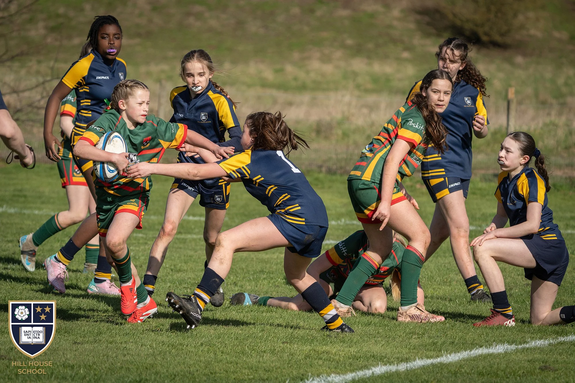 Young girls playing rugby on a grassy field, wearing colorful team uniforms with patches, some reaching for the rugby ball while others are on the ground or running. There are badges and logos on their jerseys, and the background shows a hill with gr