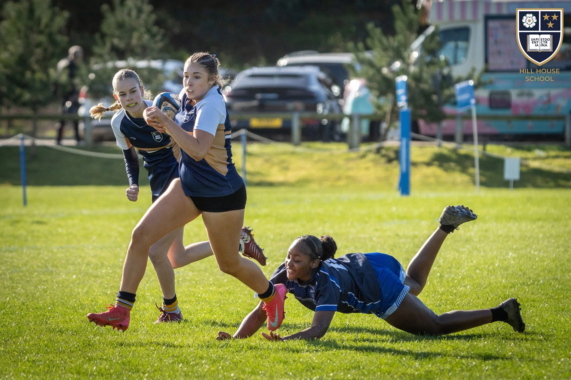 Two young girls in rugby uniforms competing for the rugby ball on a grassy field, with a third girl on the ground reaching for the ball, during daytime.