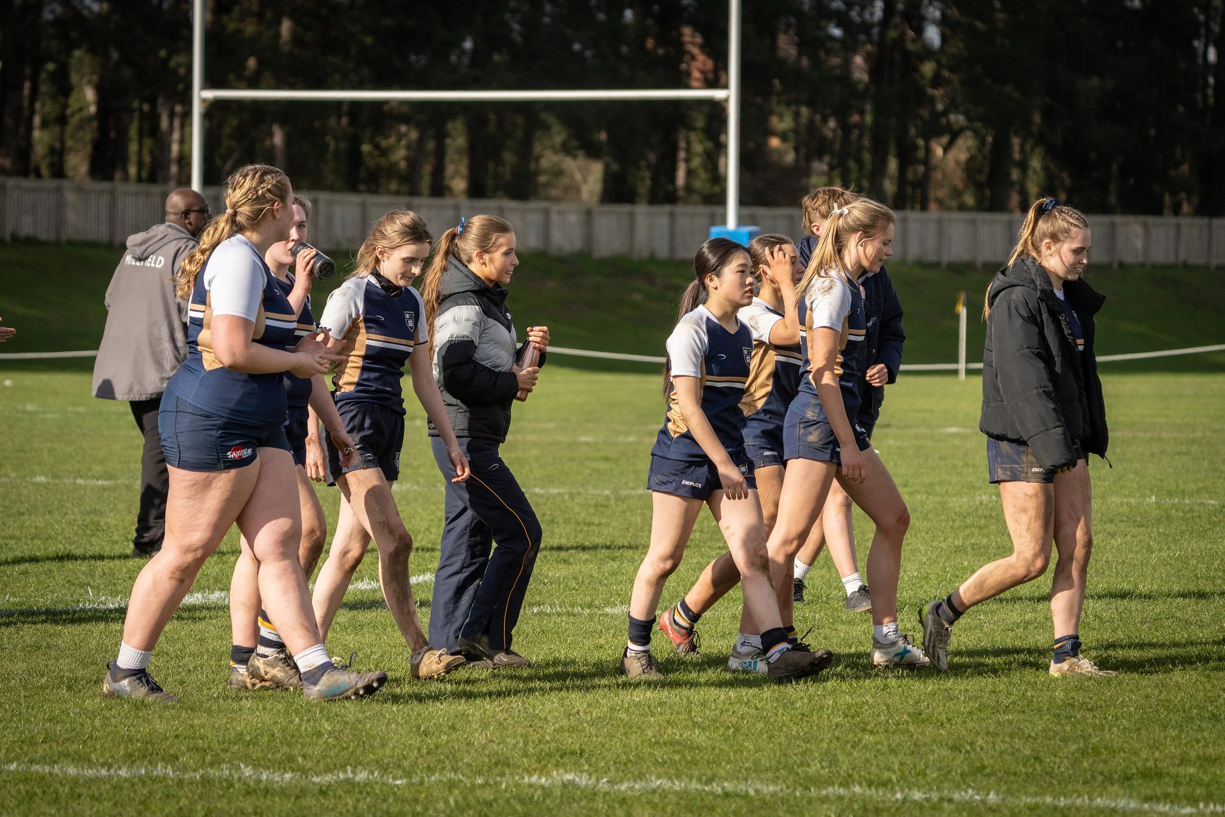 A group of young women in sports uniforms walking on a grassy field near a football goalpost.