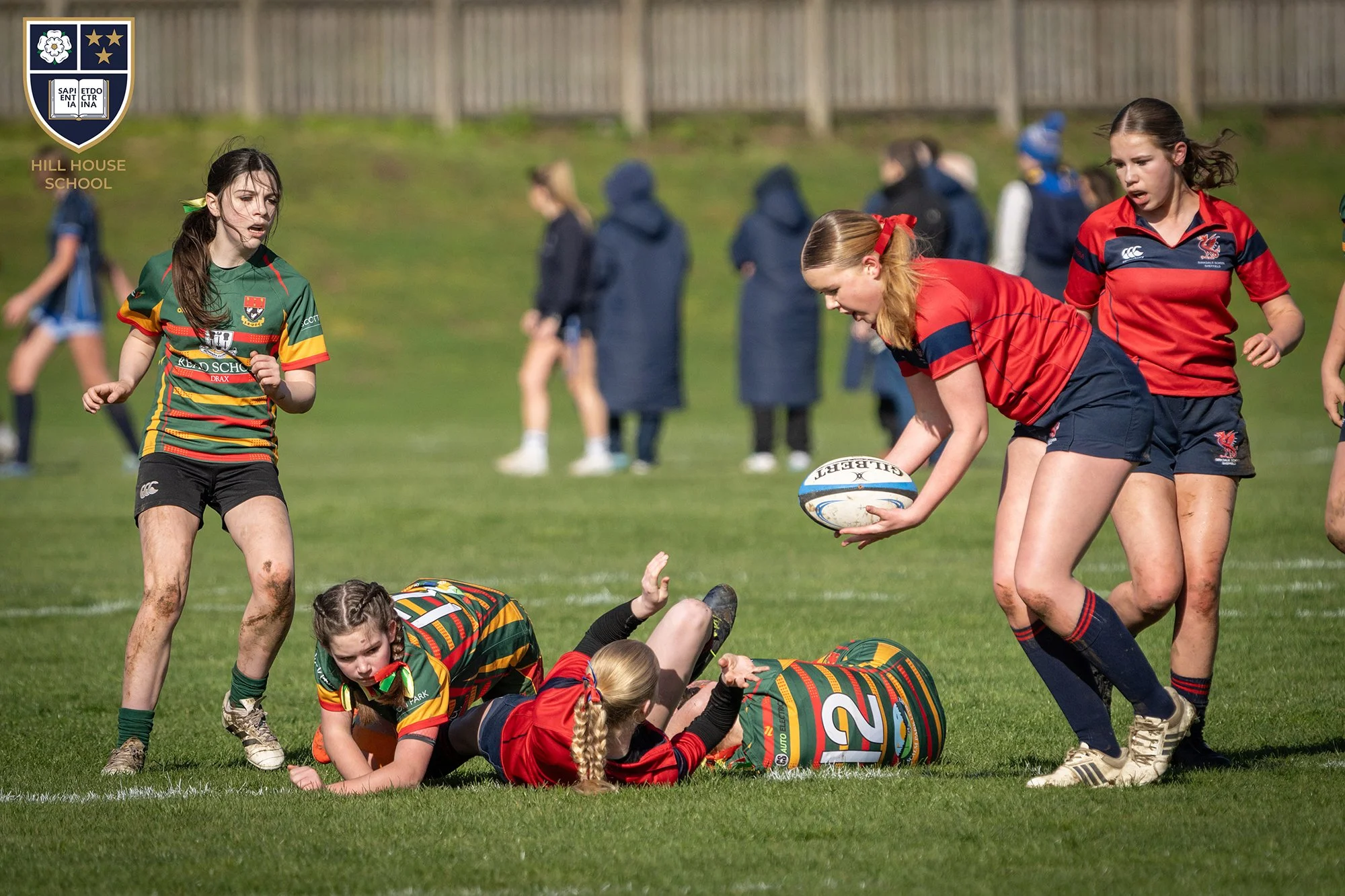 Girls playing rugby on a grassy field during daylight. One girl in green and yellow jersey stands nearby, while another in red jersey holds a rugby ball. Other players are on the ground or standing, with some spectators in coats watching in the backg